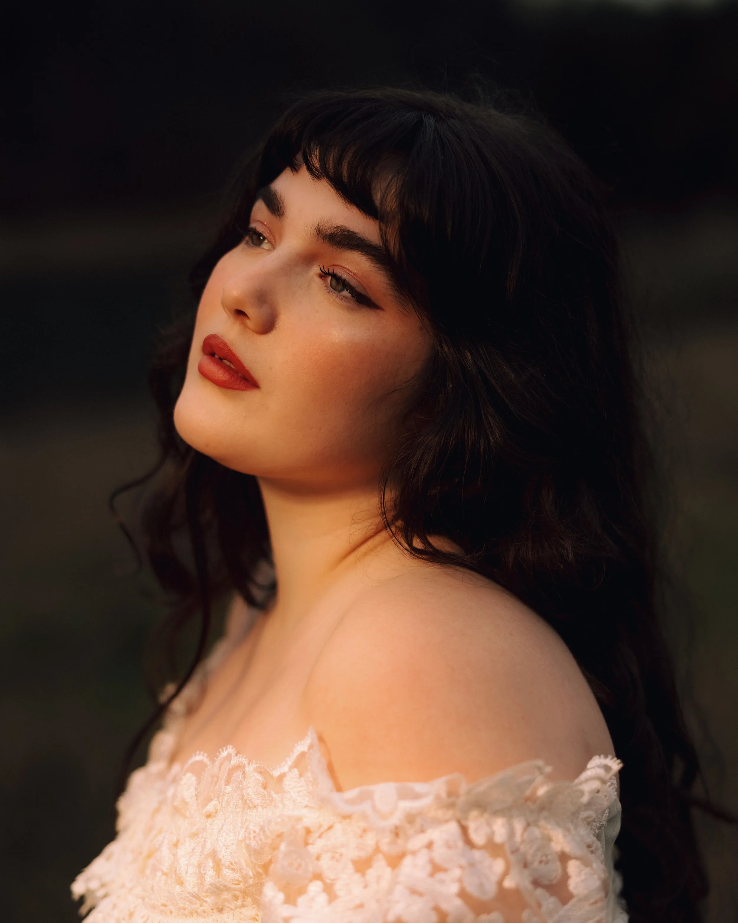 Close-up portrait of a young woman with dark hair and freckles, wearing soft makeup and a white lace dress, looking thoughtfully to the side against a dark background.