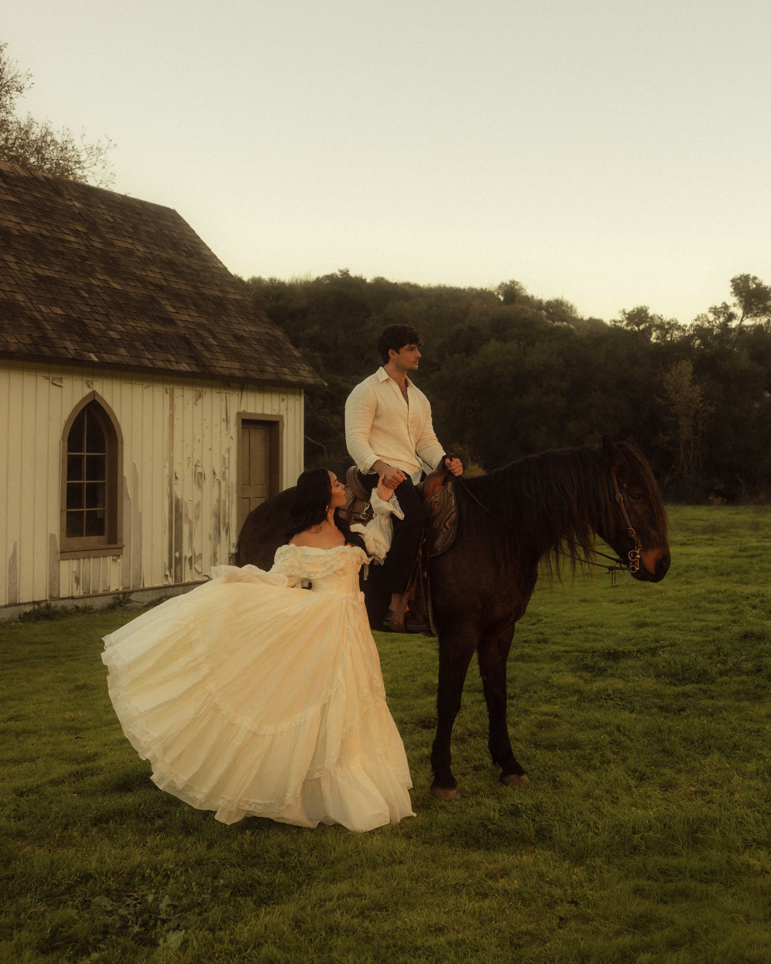 A person riding a horse on a grassy field, with two women in vintage dresses standing beside him, near a white building with arched windows and a wooden shingle roof, in a rural setting during sunset.