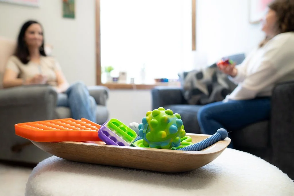 Colorful baby teething toys and a textured rubber ball on a wooden tray in the foreground, with two women sitting on couches in a living room in the background.