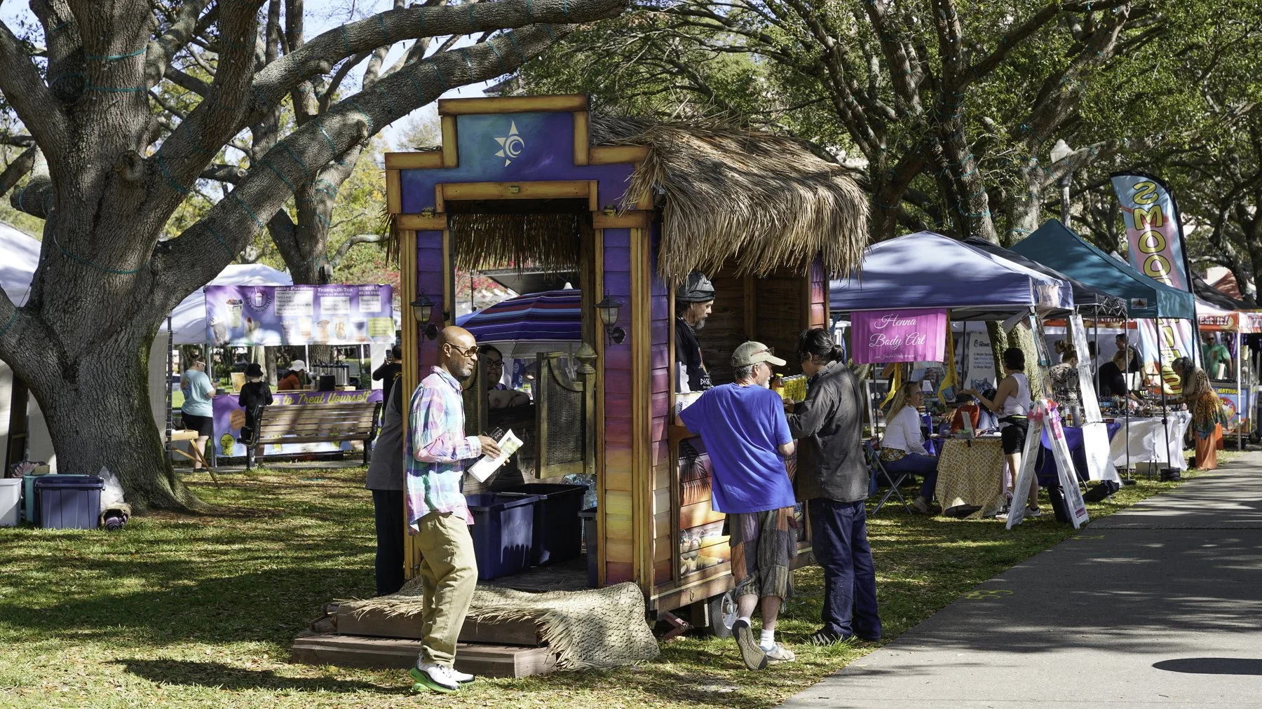 People walking around and shopping at outdoor market stalls under trees on a sunny day.
