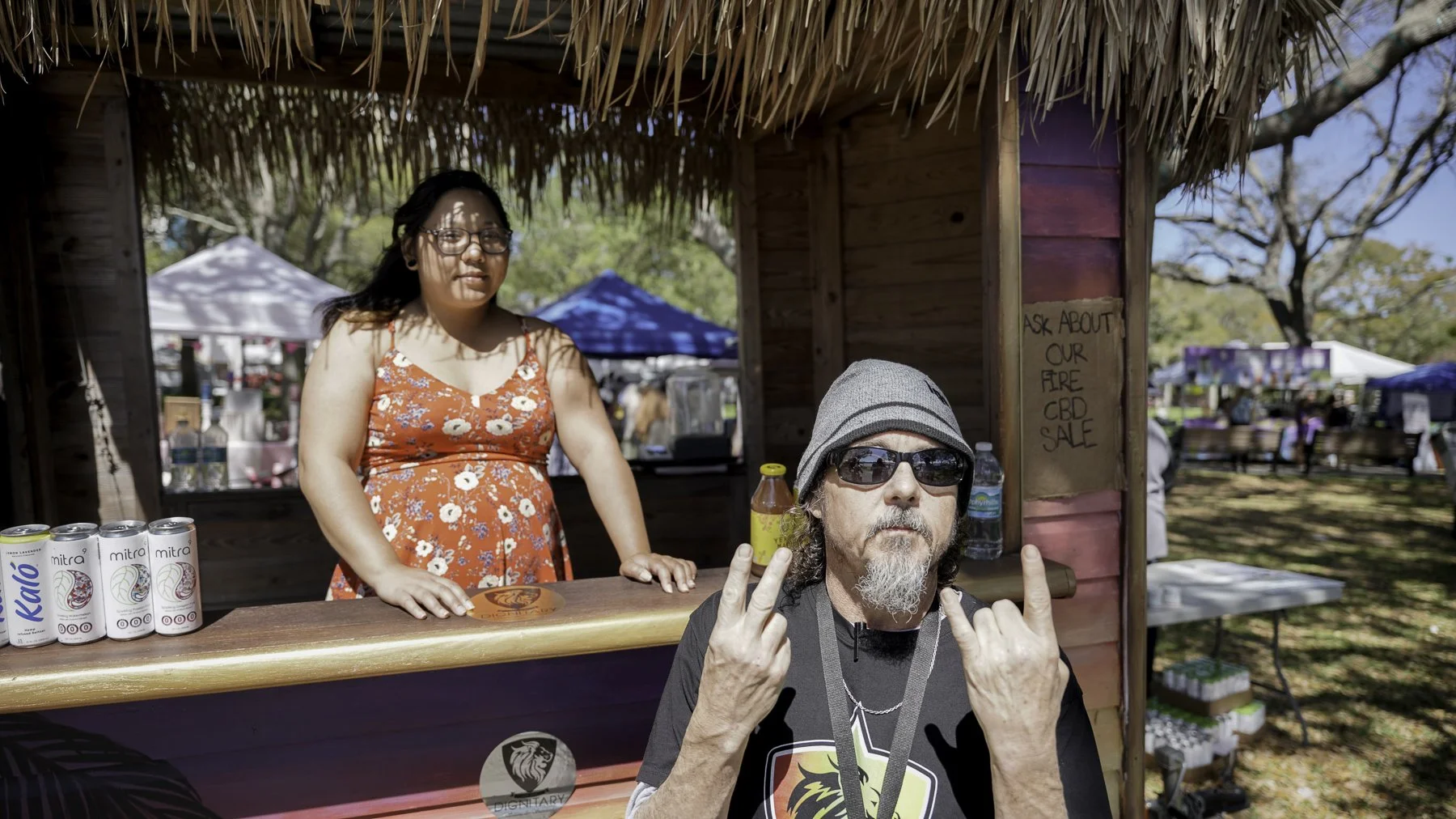 A man with sunglasses and a gray beanie making a rock and peace sign with both hands in front of a wooden booth at an outdoor market. A woman stands behind the booth wearing glasses and a red floral dress, with cans of drink on the counter. A sign on