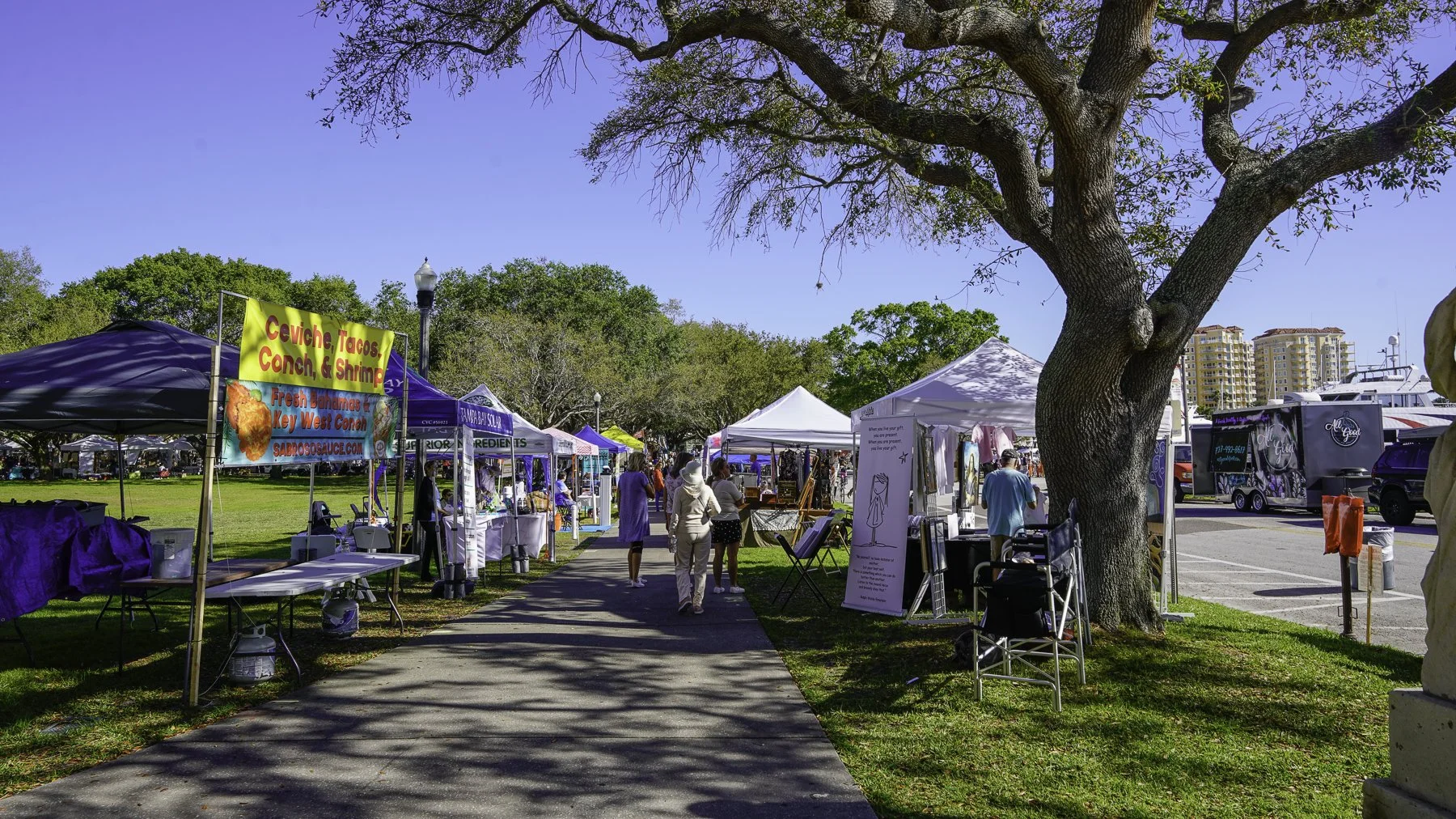 Outdoor market with vendor tents along a sidewalk, people browsing, trees, and a large tree in the foreground, RVs and buildings in the background.