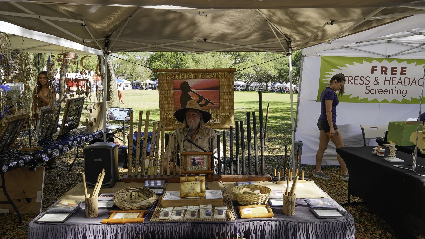 Vendor at outdoor craft market booth displaying bamboo flutes and other handmade items with woman wearing a wide-brimmed hat, background includes jewelry displays, another woman walking past, and a banner offering free stress and headache screenings.