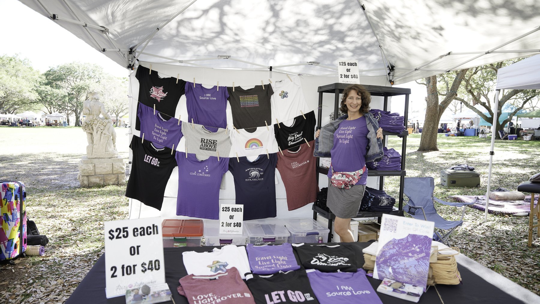 A woman standing in a market stall displaying various colorful T-shirts hanging on a line, with signs showing prices of $25 each or 2 for $40. The stall has shelves with more T-shirts, and a table with T-shirts folded. The market is outdoors with tre