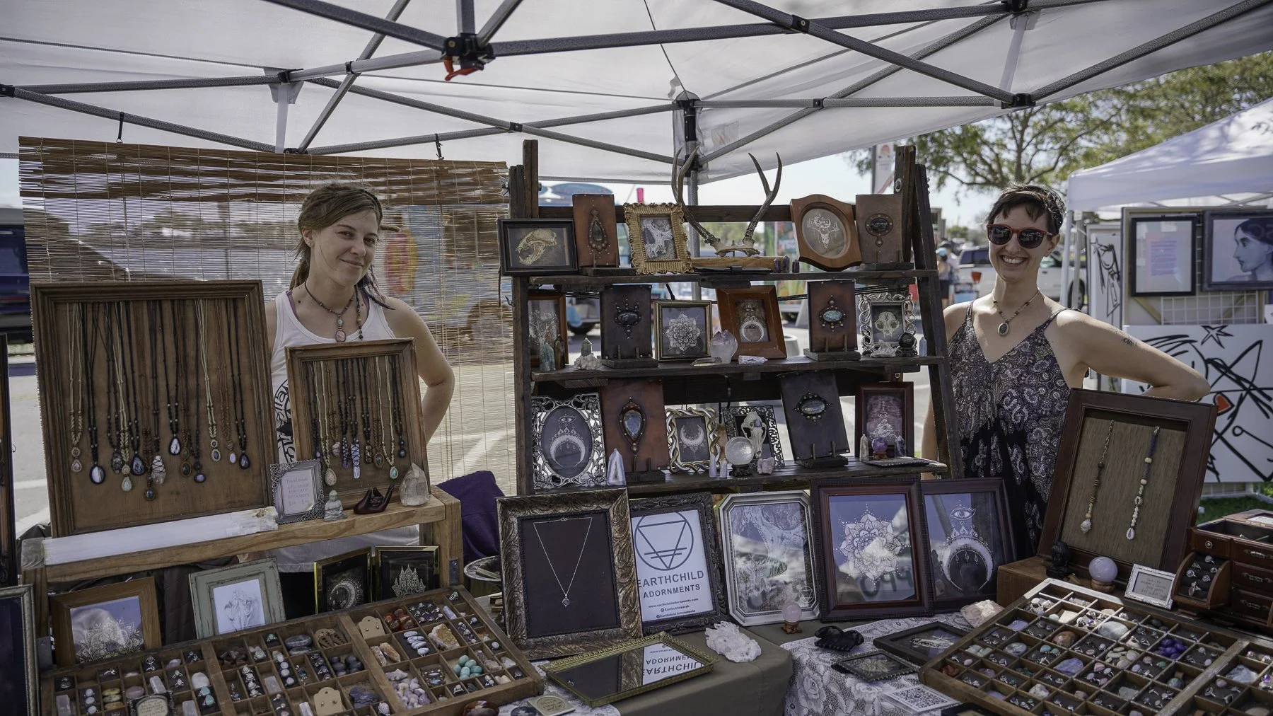 Two women at a jewelry stall outdoors, displaying necklaces, rings, and decorative items under a tent.