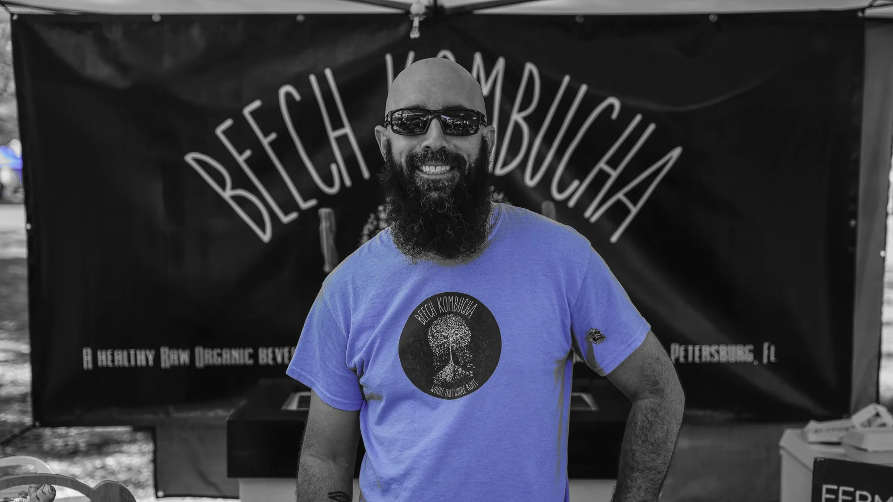 A man with a beard and bald head wearing sunglasses and a blue T-shirt standing in front of a black stall banner that reads "BEACH KOMBUCHA". The T-shirt has a logo with a tree and the text "Whole Fruit Whole Root." The man is smiling with his arm on