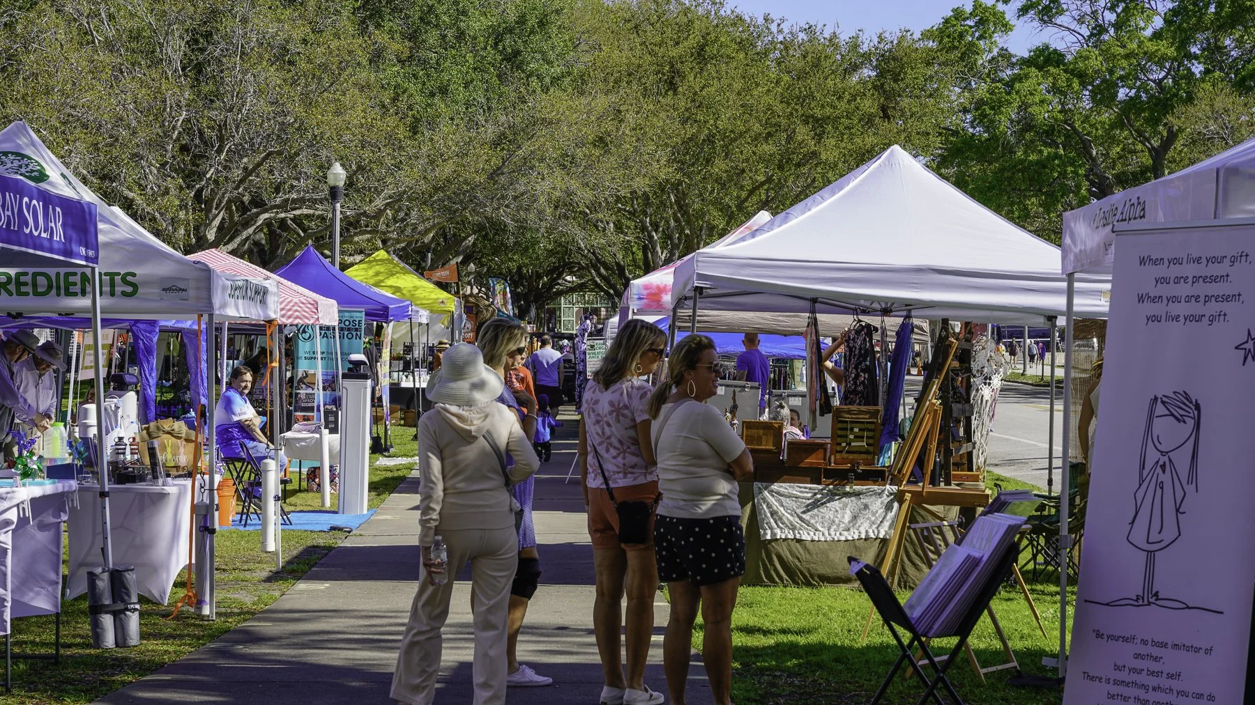 People browsing outdoor market stalls with tents and trees in background.