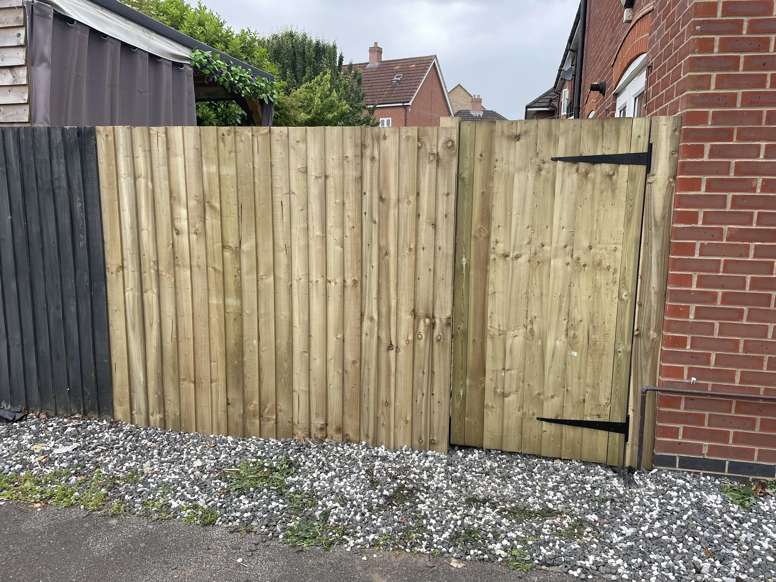 A wooden backyard fence with a gate, painted in natural wood with black hinges, next to a brick house with a white window, on a gravel ground with some grass and weeds.