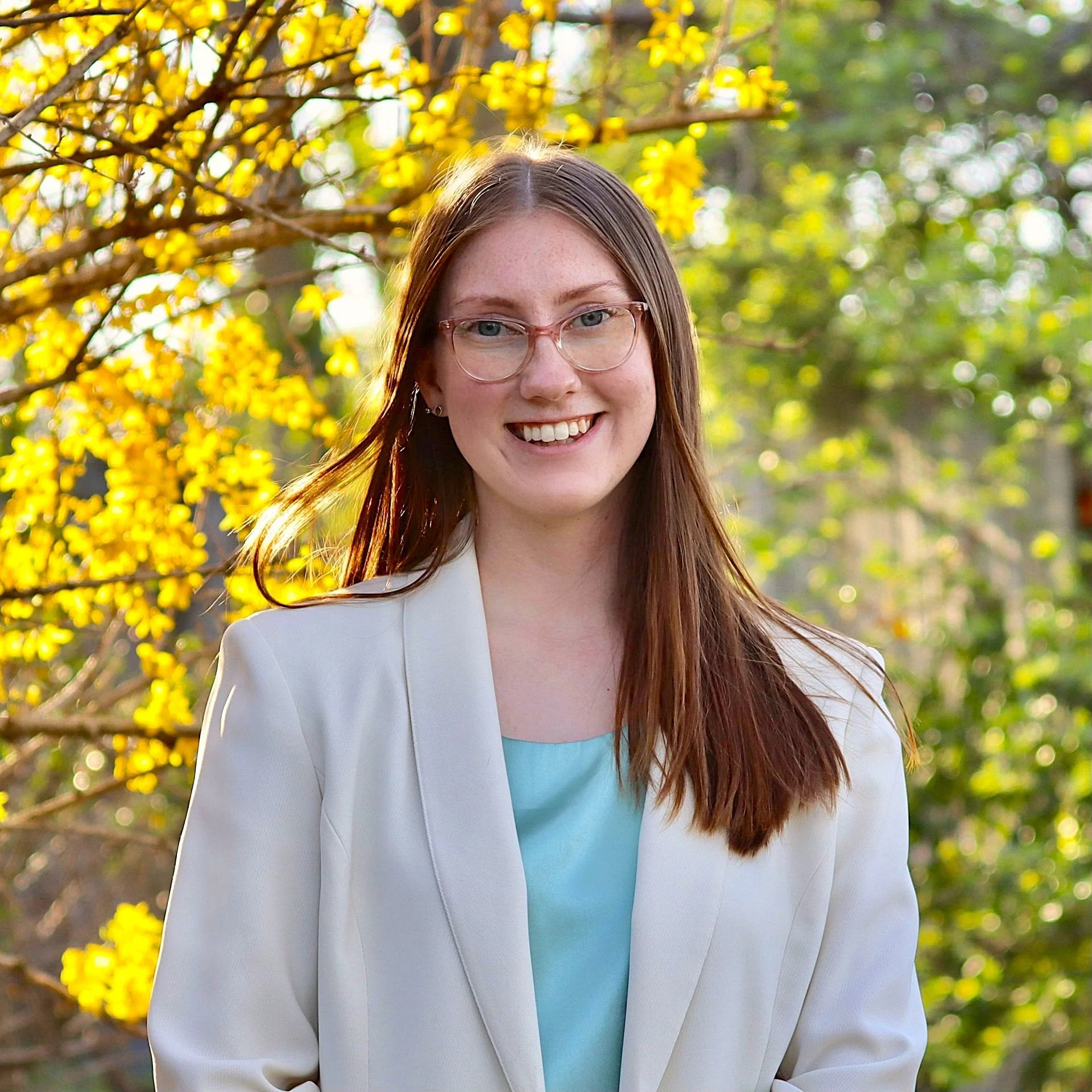 A young woman with long red hair, glasses, and a white blazer smiling outdoors in front of yellow flowering bush with green foliage.
