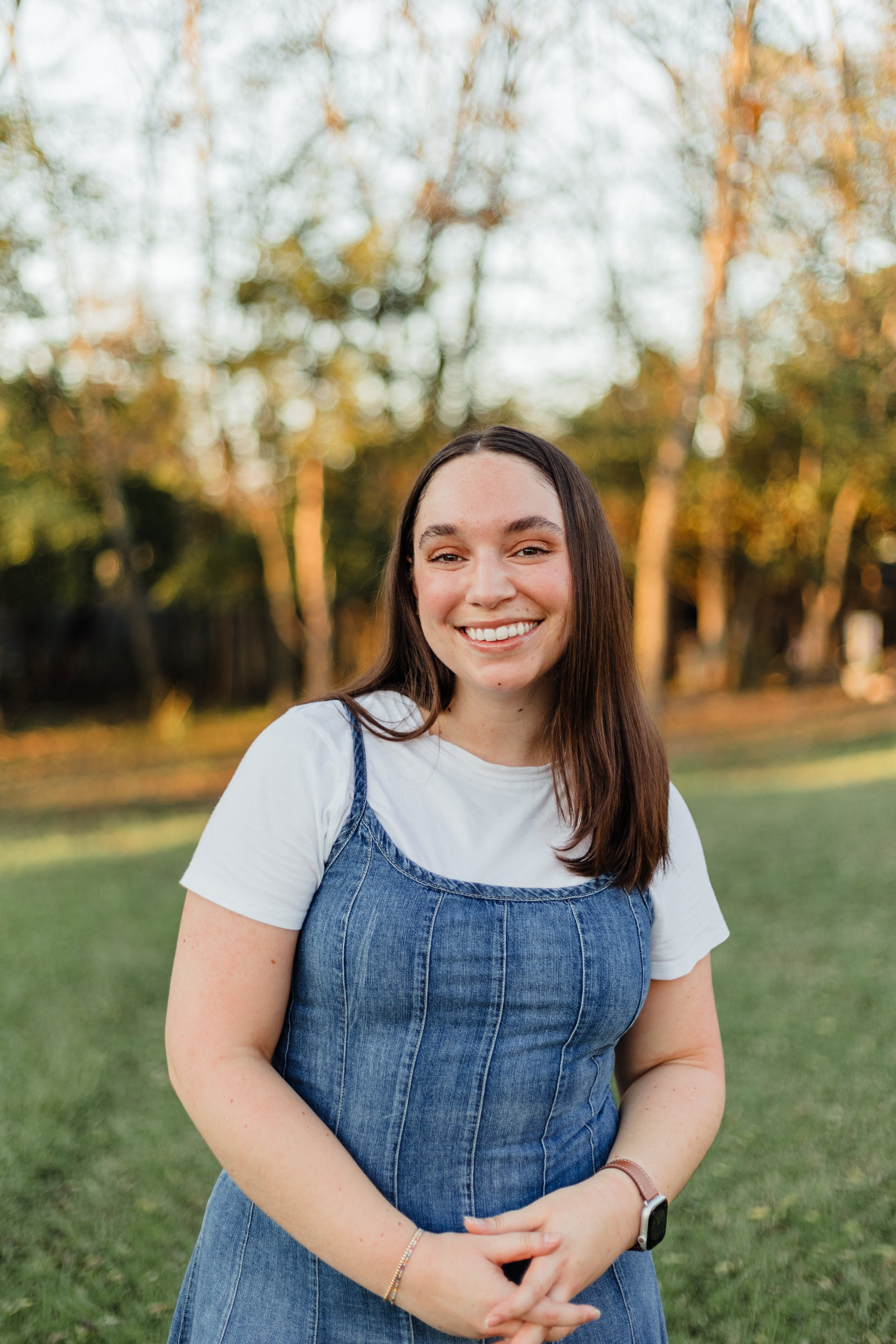 A woman with long dark hair smiling outdoors during sunset, wearing a white t-shirt and a denim dress, standing on grass with trees in the background.
