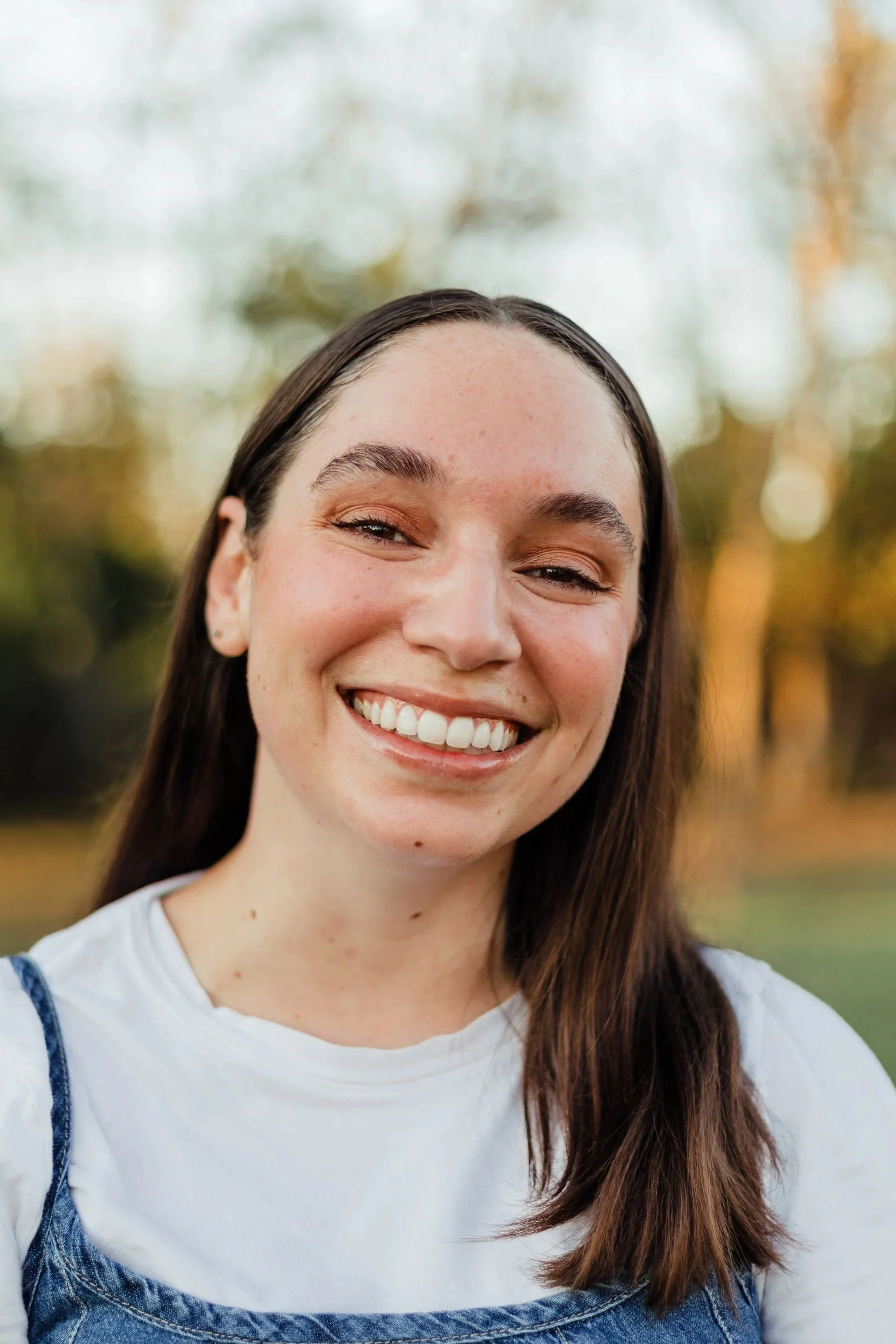 A young woman with long brown hair smiling outdoors during fall, wearing a white t-shirt and denim overalls.