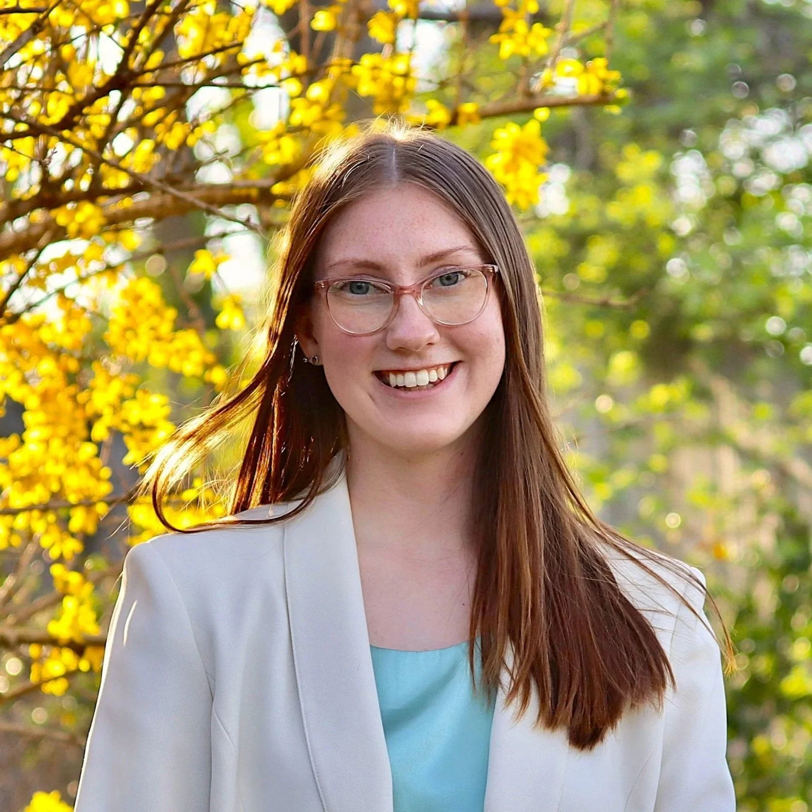 A smiling woman with long red hair and glasses, wearing a white blazer over a light blue top, standing outdoors with yellow flowers and green leaves in the background.