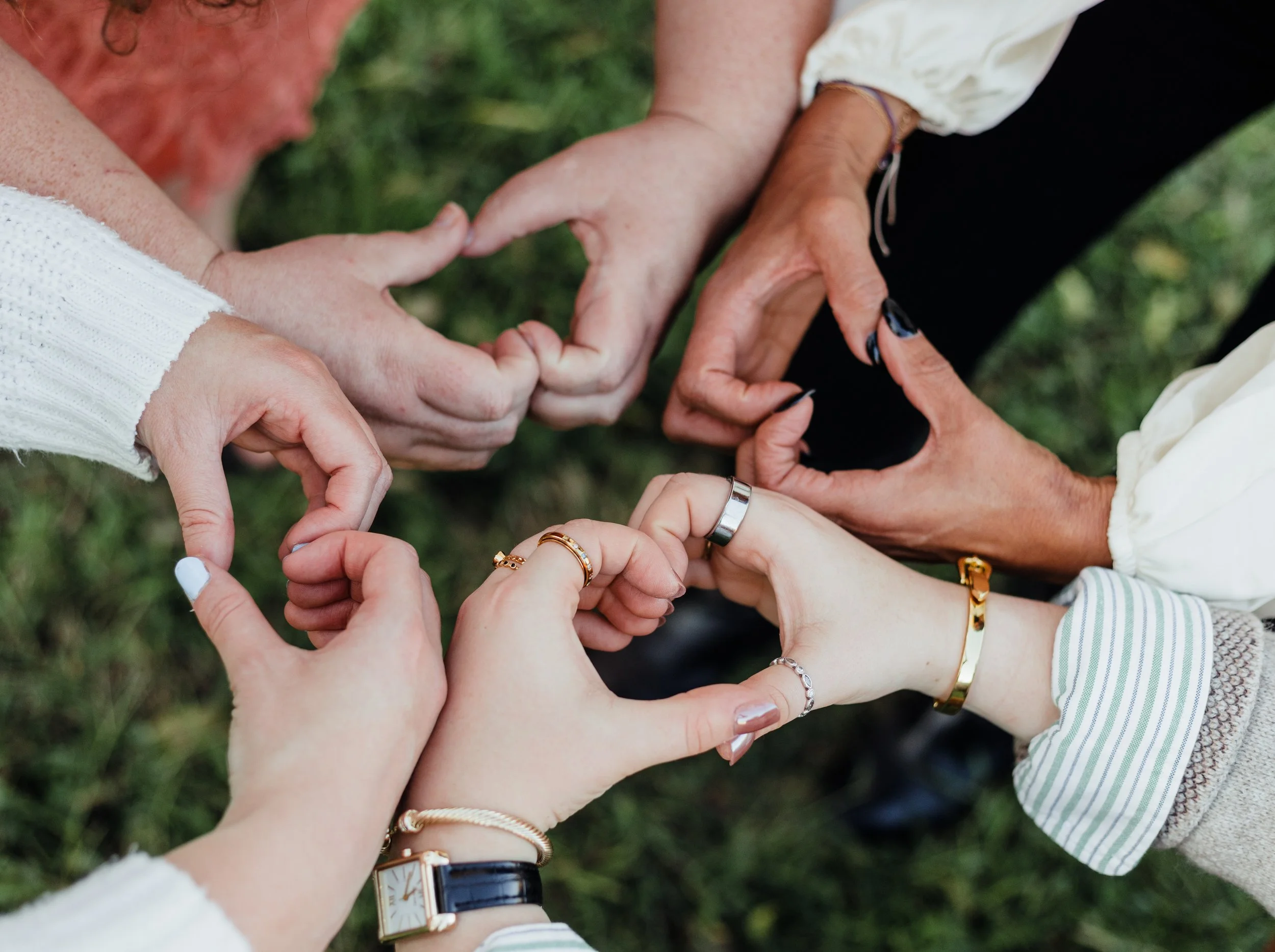 Multiple hands forming heart shapes with fingers, displaying rings and bracelets, outdoors on grass.