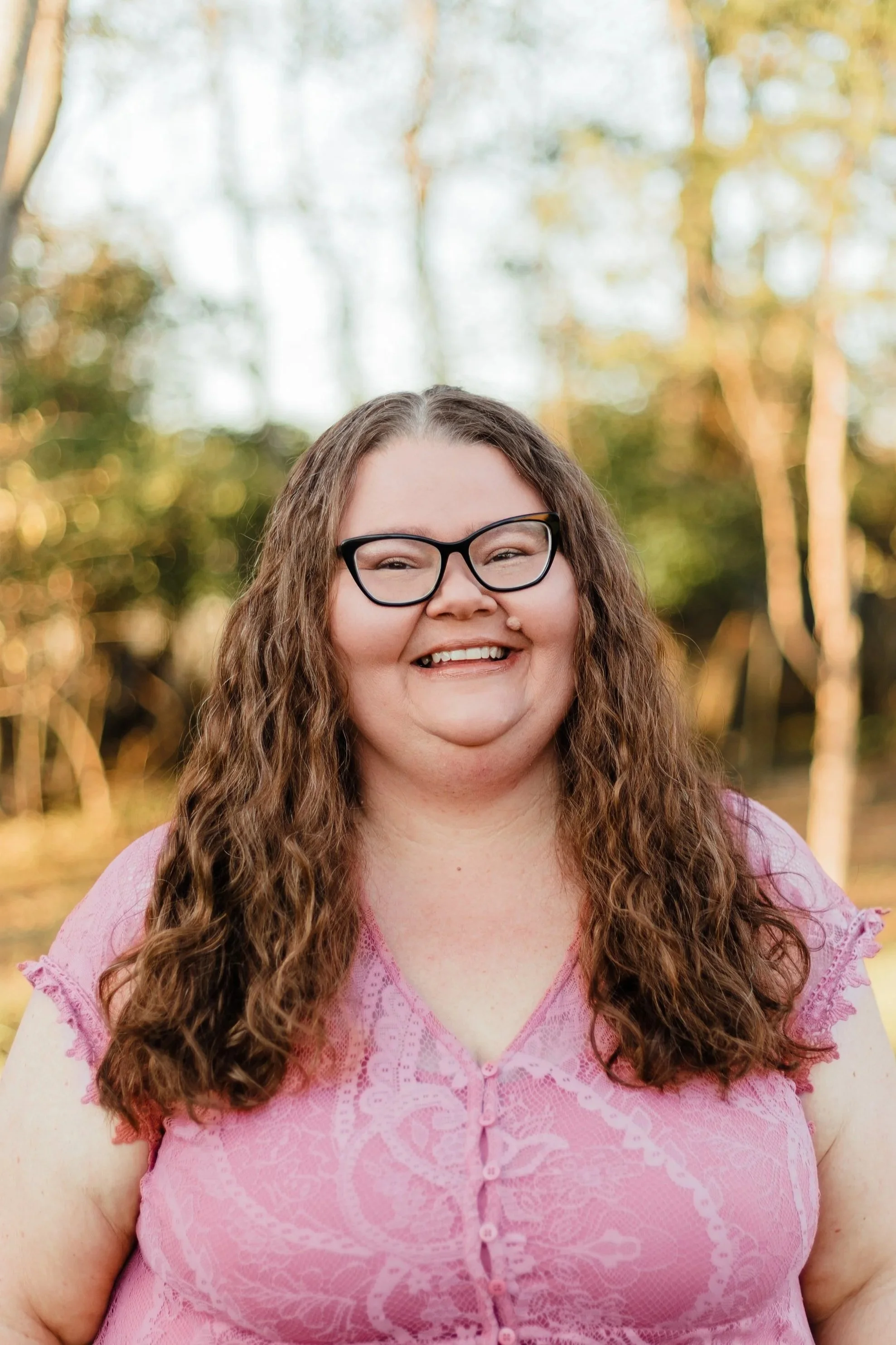 A woman with long curly hair, glasses, and a pink lace blouse is smiling outdoors with trees and sunlight in the background.