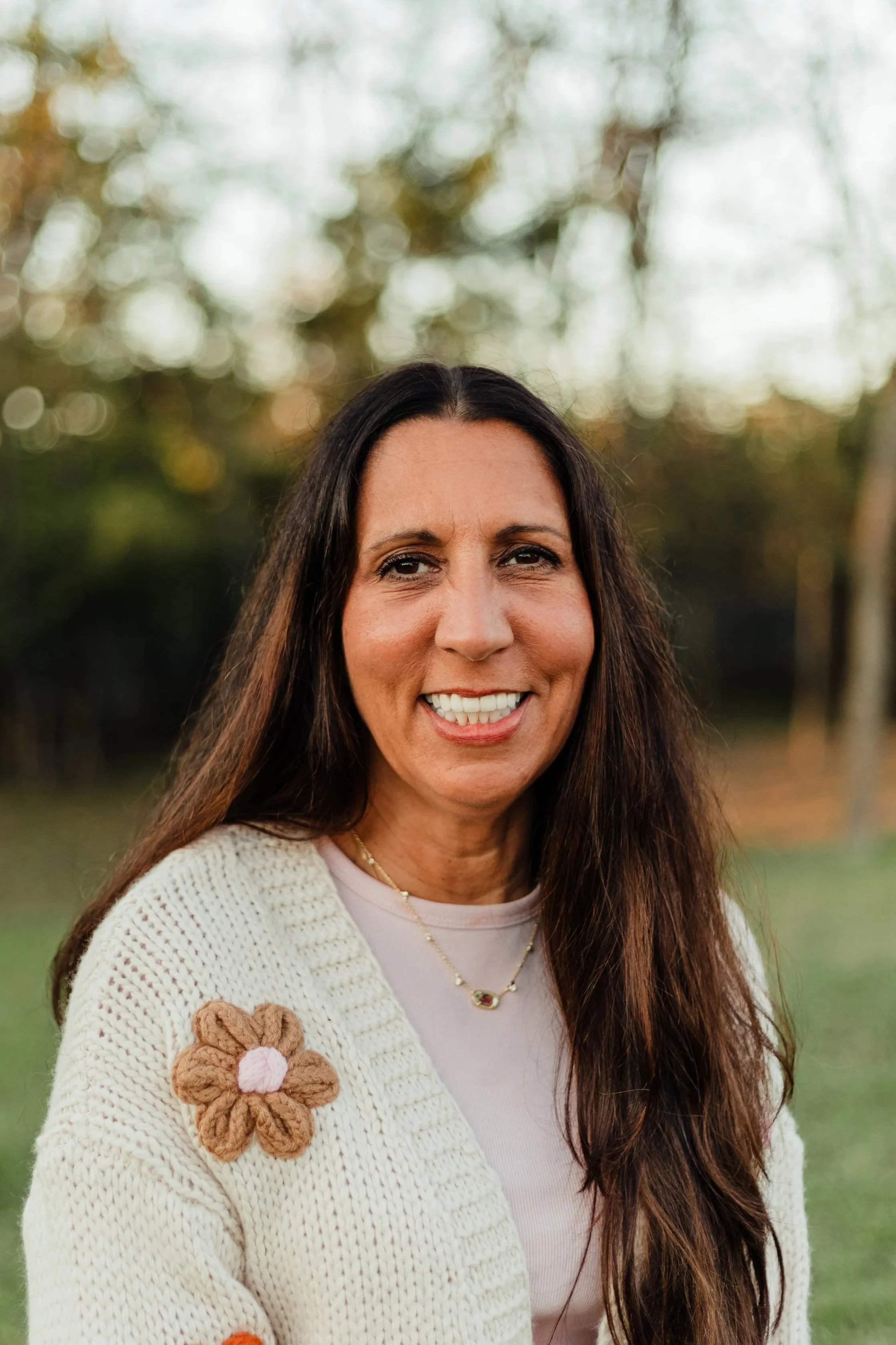 A woman smiling outdoors with trees in the background, wearing a cream-colored sweater with flower embellishments and a necklace.