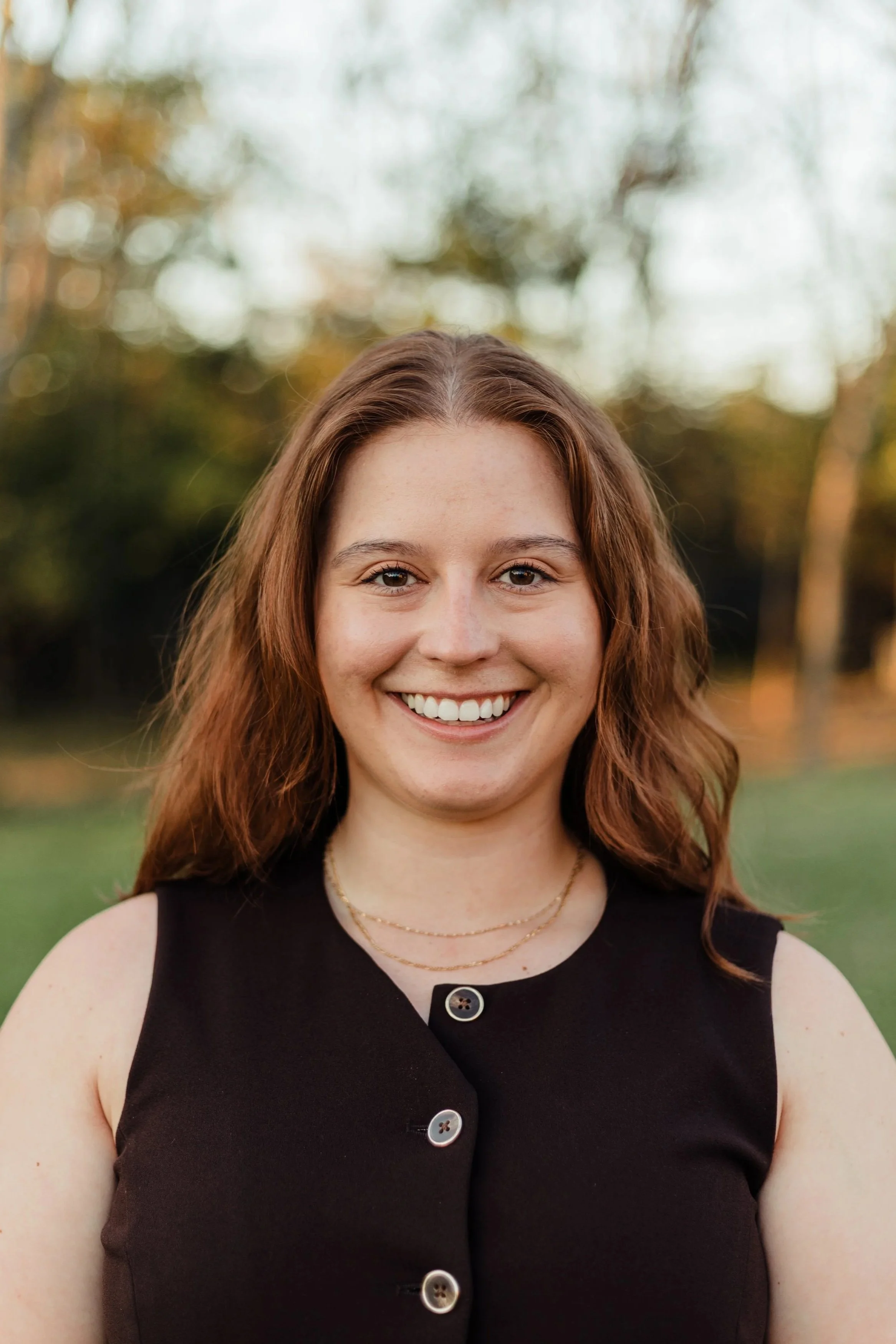 A young woman with wavy auburn hair smiling outdoors in a park setting with trees and blurred background.