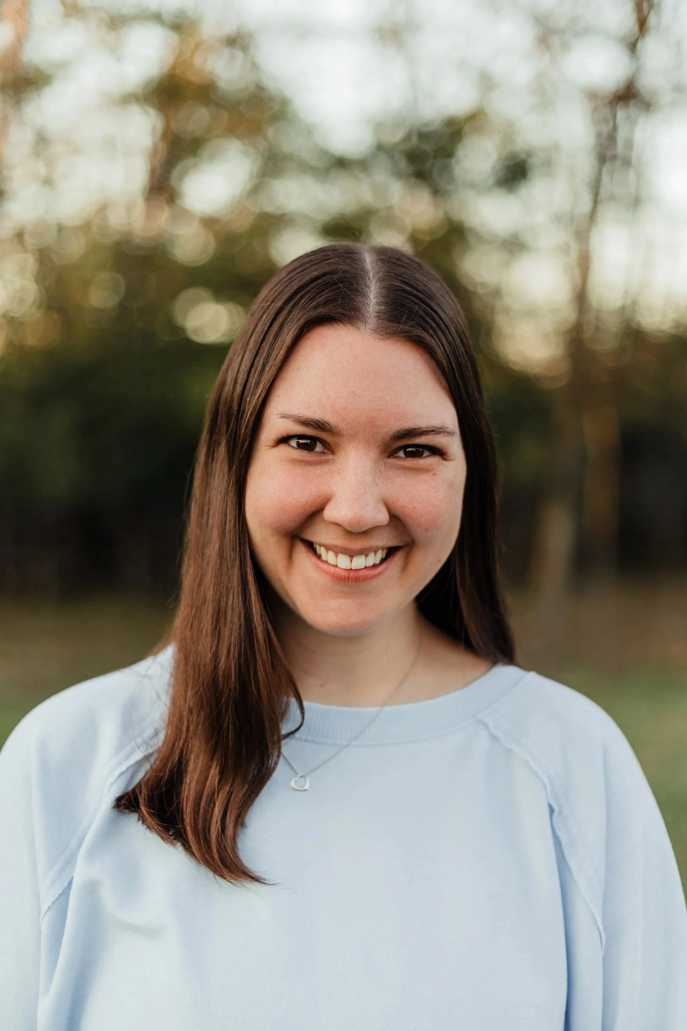 A young woman with long brown hair smiling outdoors, wearing a light blue sweatshirt and a necklace with a heart-shaped pendant, with trees in the background.