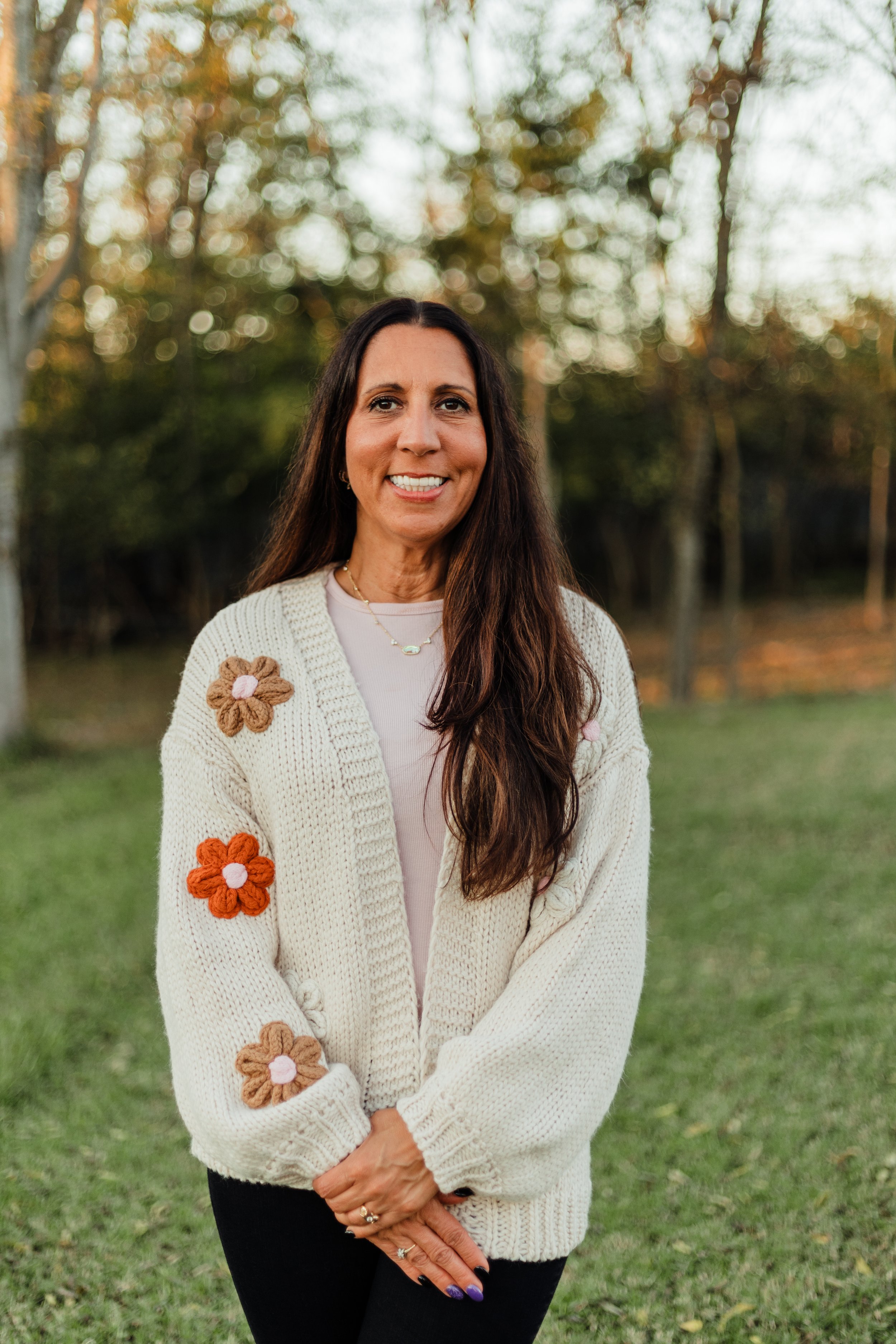A woman with long brown hair smiling outdoors in a park during fall, wearing a cream sweater with flower patches and a light-colored shirt underneath.