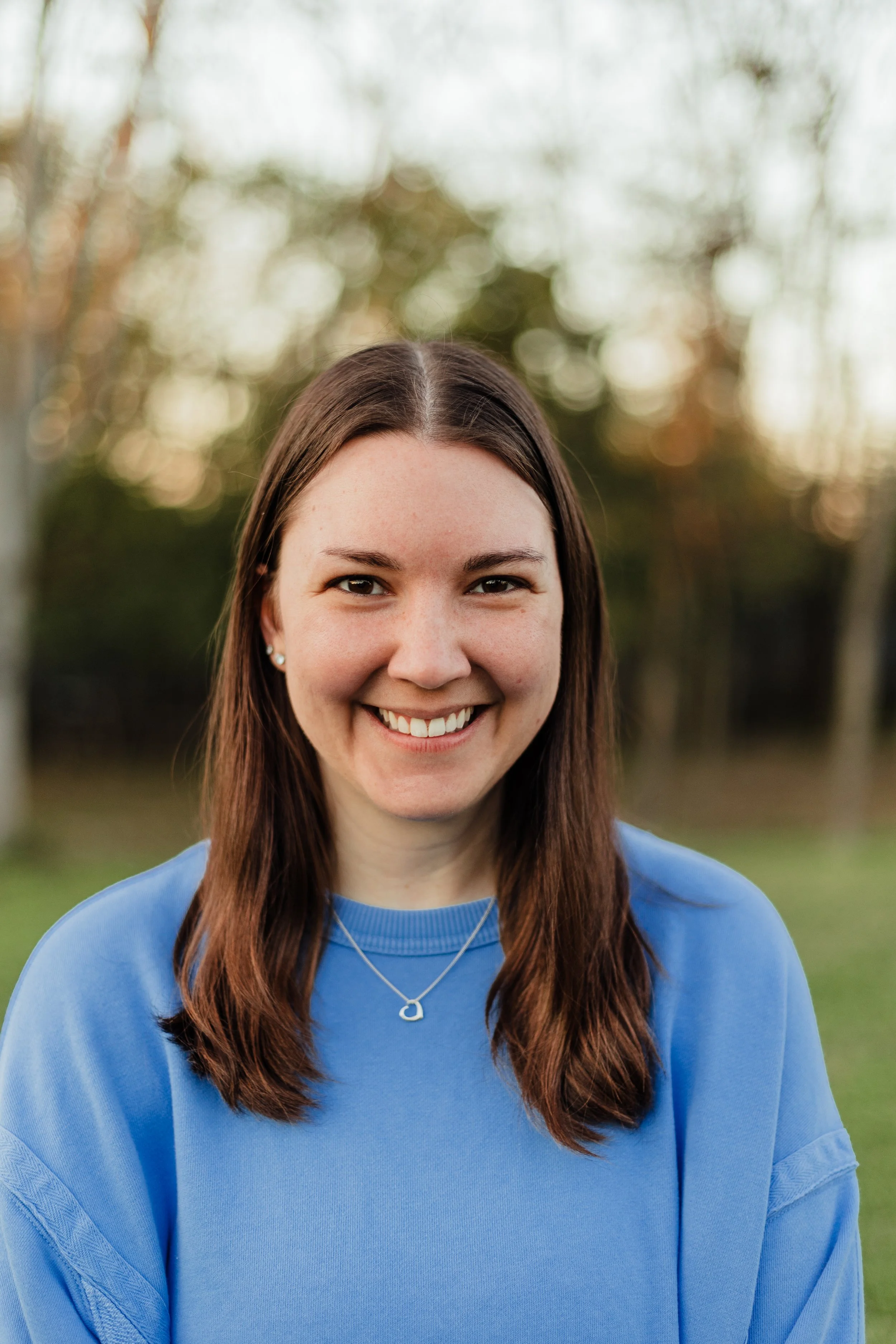 A young woman with brown hair and a blue sweatshirt, smiling outdoors with trees in the background during sunset.