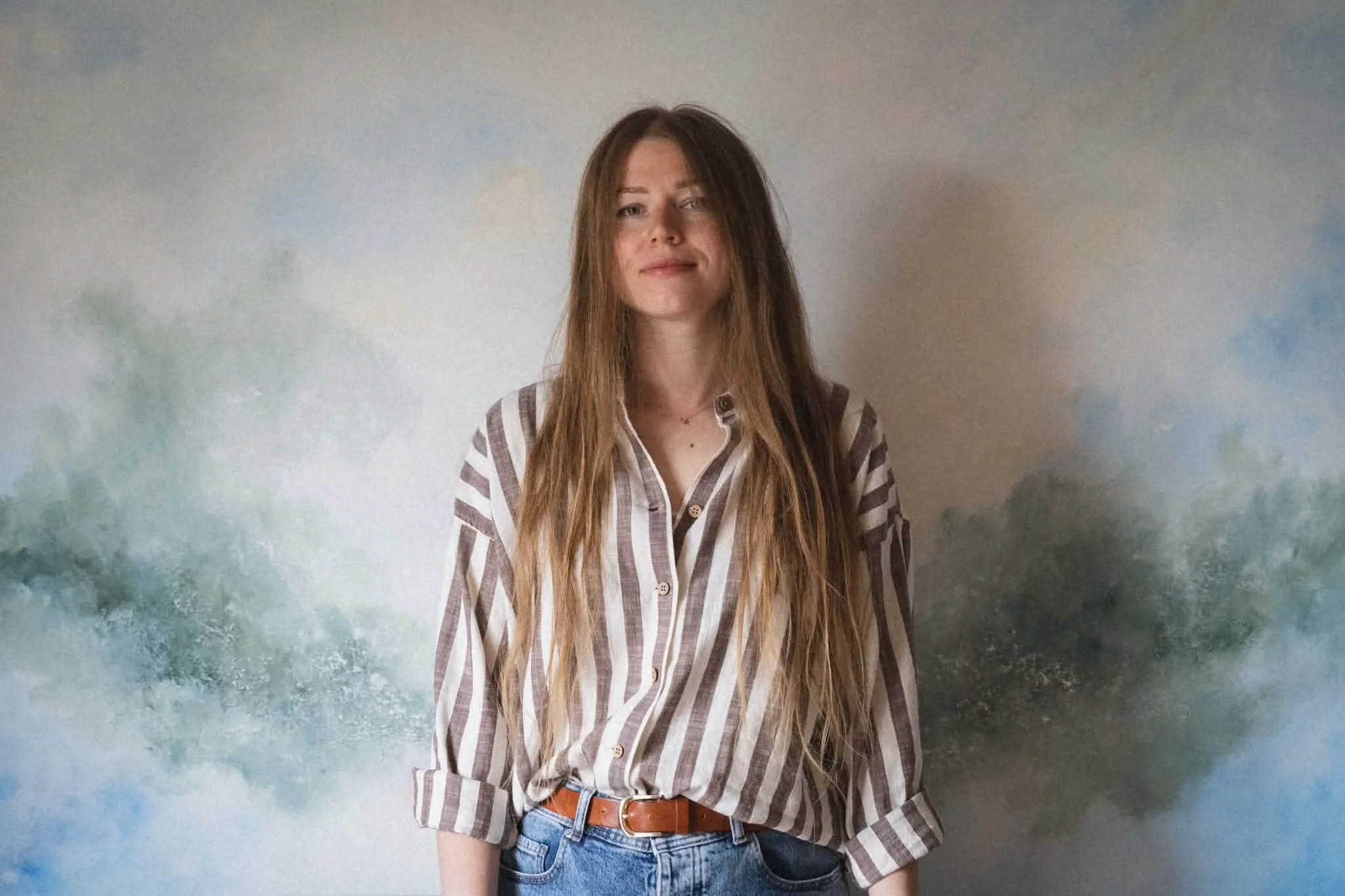 A young woman with long, wavy hair standing in front of a painted sky mural background, wearing a brown and white striped shirt with sleeves rolled up, tucked into high-waisted jeans with a brown belt.