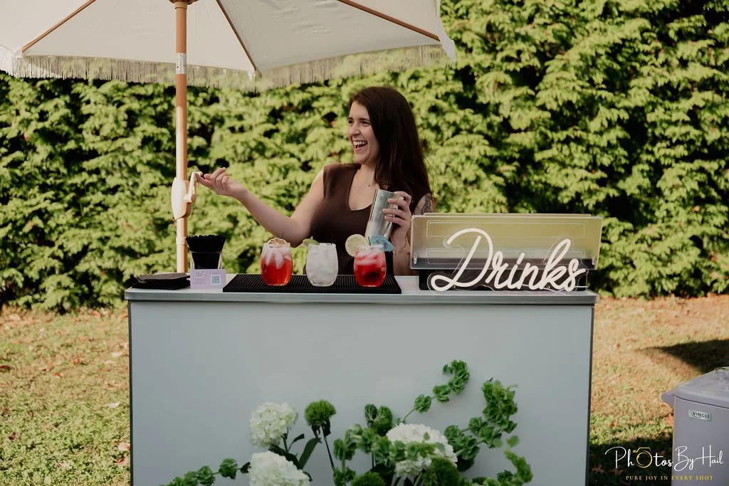 the owner, Cara, laughing behind the mobile bar set up with a drink shaker in hand.