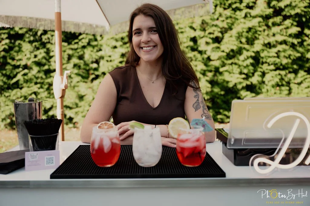 the owner, Cara, smiling behind the bar with a row of cocktails made in front of her