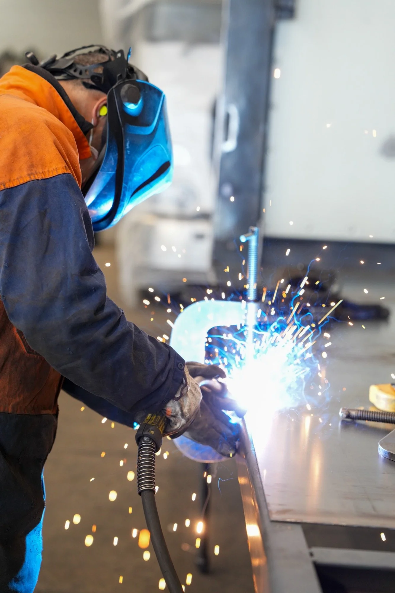 A person welding metal at a Fruehauf worktable, wearing protective gear including gloves, a face shield, and ear protection, with bright sparks flying from the welding process.