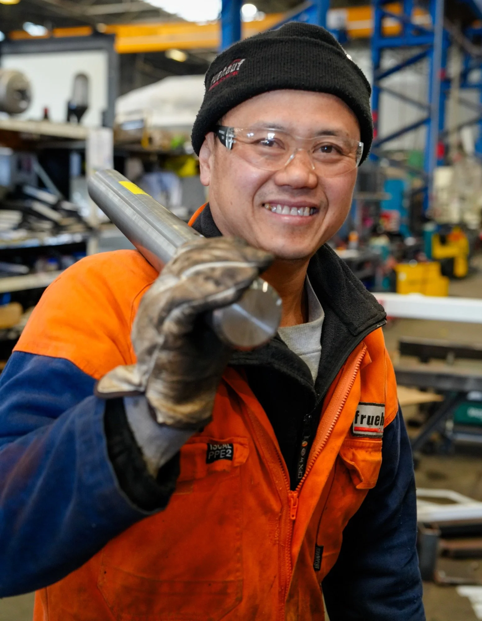 A smiling man in safety glasses, black beanie, orange and blue work jacket, and gloves, holding a metal pipe in a Fruehauf truck and trailer workshop with various tools and equipment in the background.