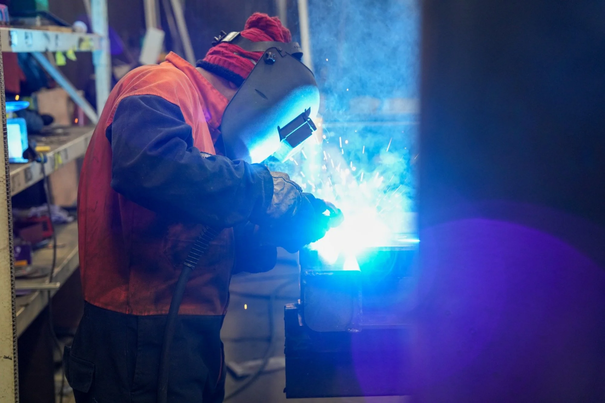 Person welding in a Fruehauf truck and trailer workshop, wearing a welding helmet and protective clothing, sparks and bright blue light from the welding process.