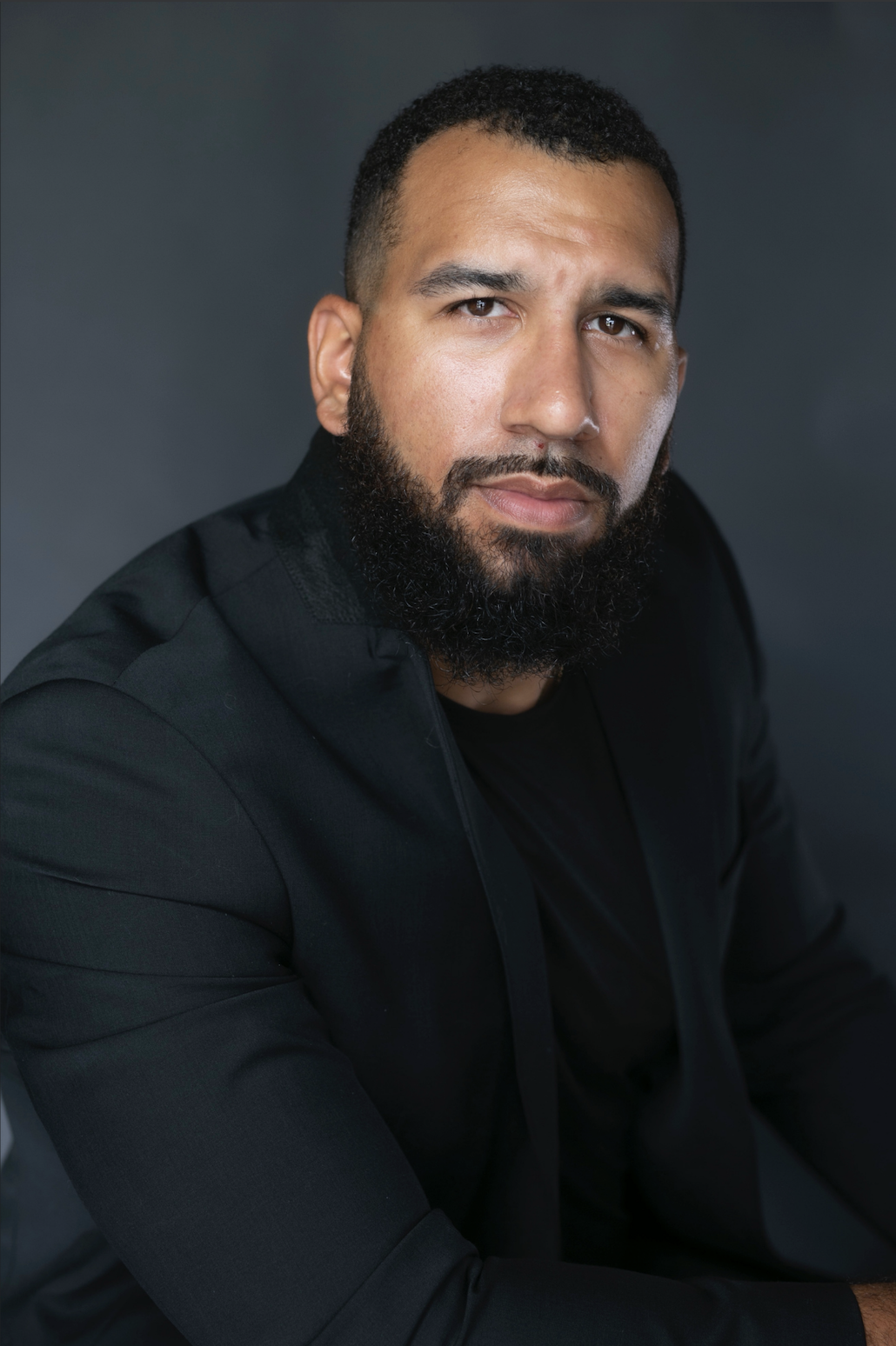 A man with a beard and short hair wearing a black jacket and shirt, looking at the camera against a dark background.