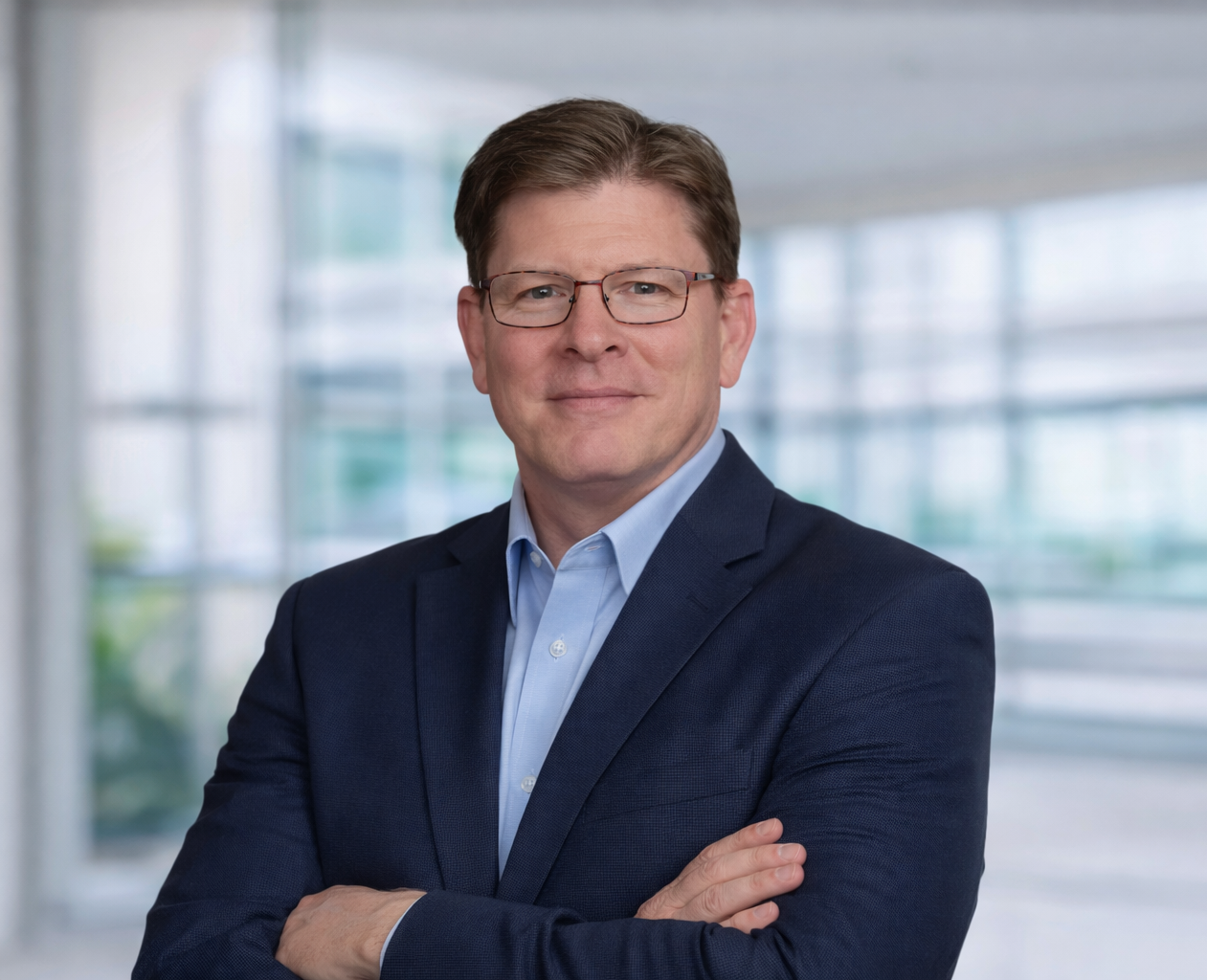A professional man with short brown hair, glasses, wearing a navy blue suit and light blue dress shirt, standing with arms crossed in front of a modern office building with large glass windows.