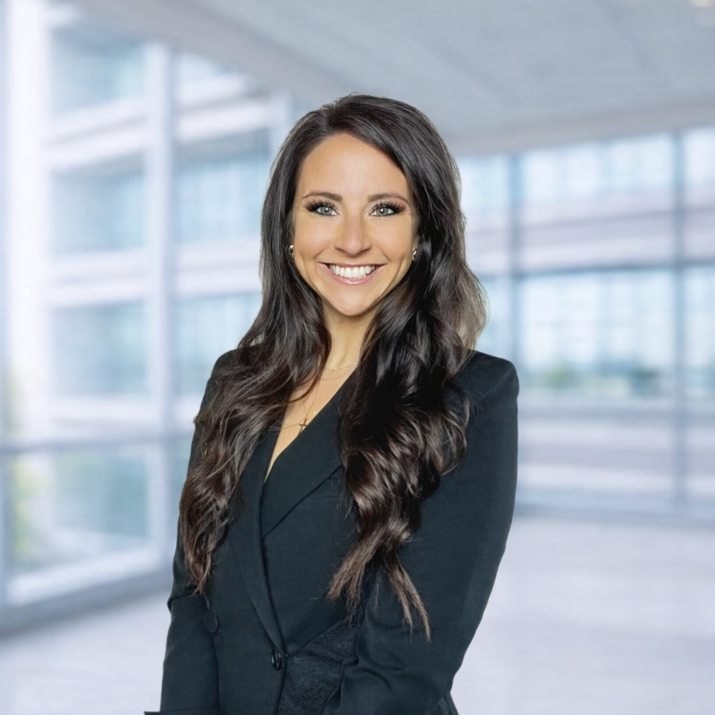 A smiling woman with long dark hair dressed in a black blazer standing in a modern office building with large windows.
