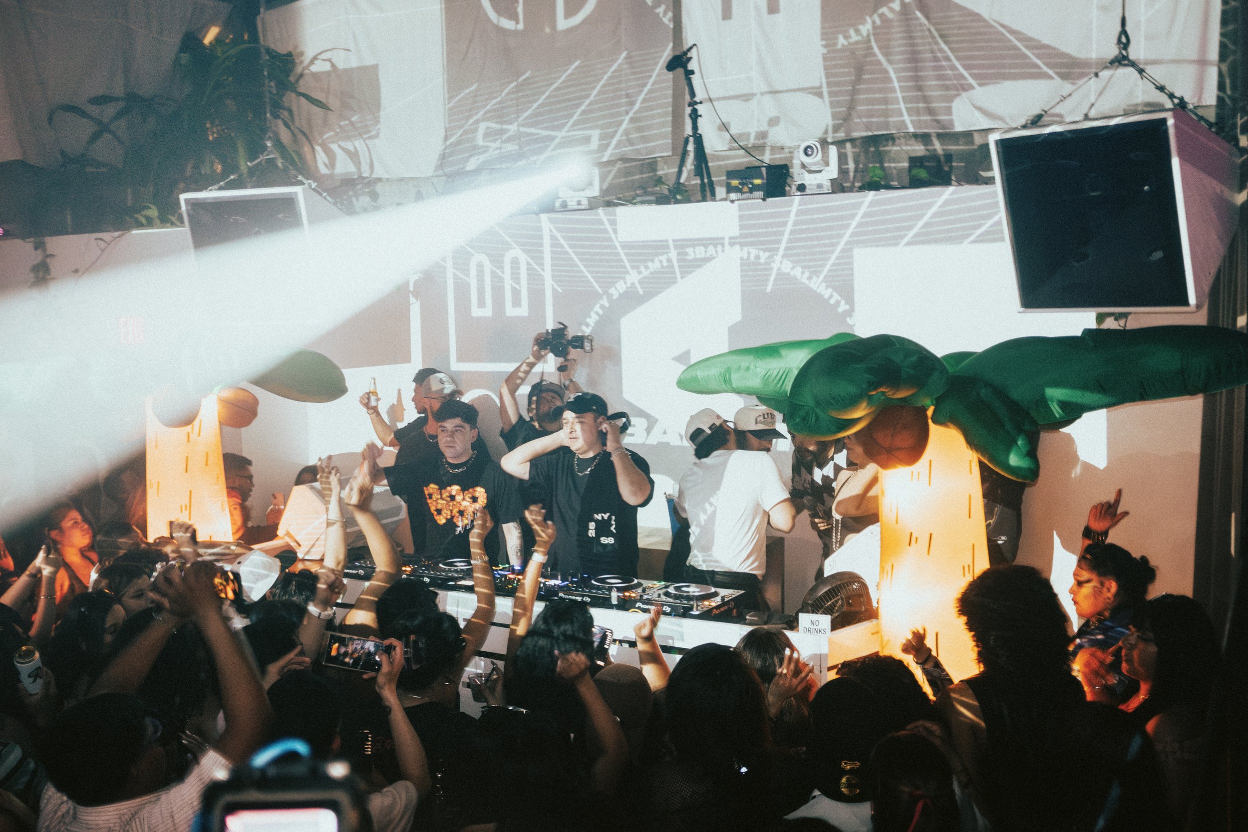 A lively party scene with a DJ performing on stage, surrounded by enthusiastic attendees dancing and taking photos. The venue has vibrant lighting, large decorative inflatable props, and a projected backdrop with graphics and text.