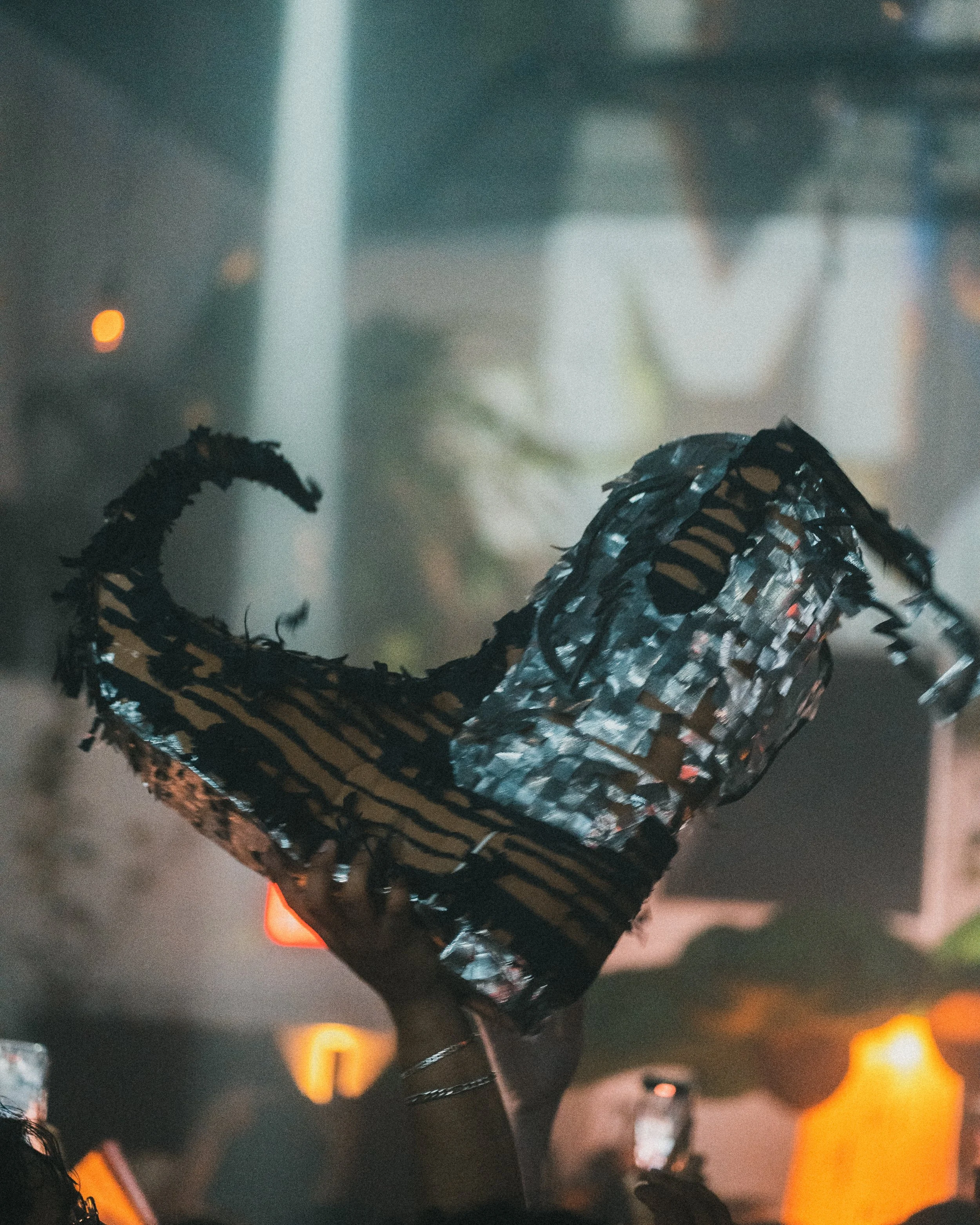 A person is holding up a painted pinata of a bota  in a crowded, dimly lit indoor space with blurred background lights.
