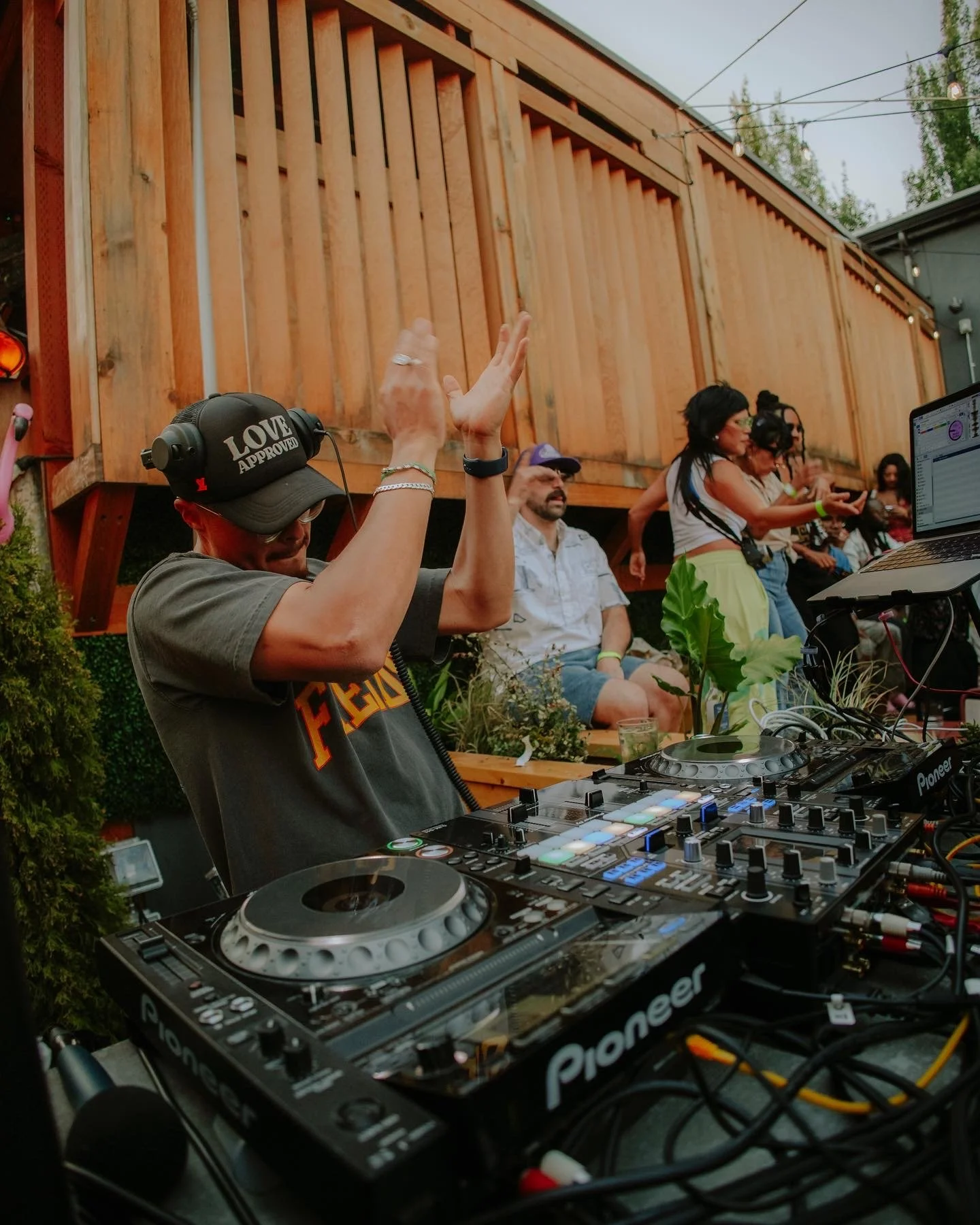A DJ wearing a black cap with 'LOVE APPROVED' written on it, headphones, and a gray T-shirt is dancing with his hands raised in the air at an outdoor event with a wooden fence and a group of people in the background.