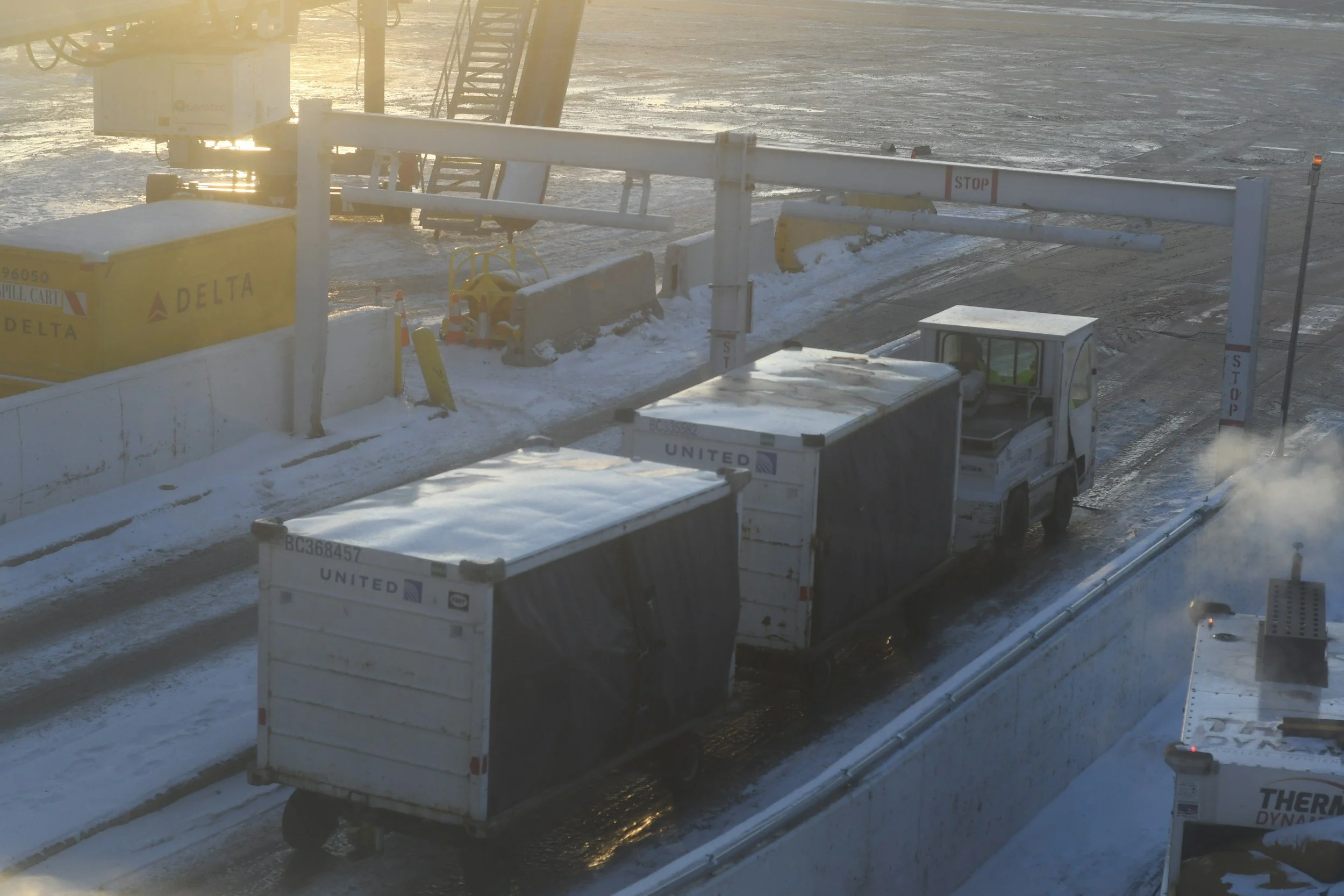 Two trucks are parked at a snowy toll booth on a winter day, with a barrier arm and caution signs, and snow-covered ground and a sunset in the background.