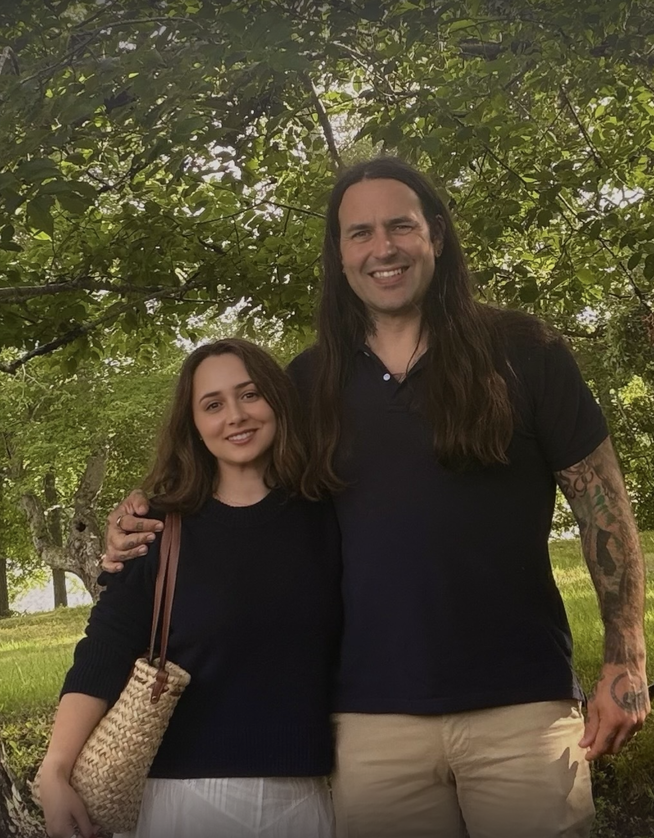 A young woman and a man standing together outdoors under a tree, smiling at the camera. The woman has shoulder-length brown hair and wears a black sweater, holding a woven bag. The man has long dark hair, visible tattoos on his arms, and wears a black polo shirt and beige shorts.