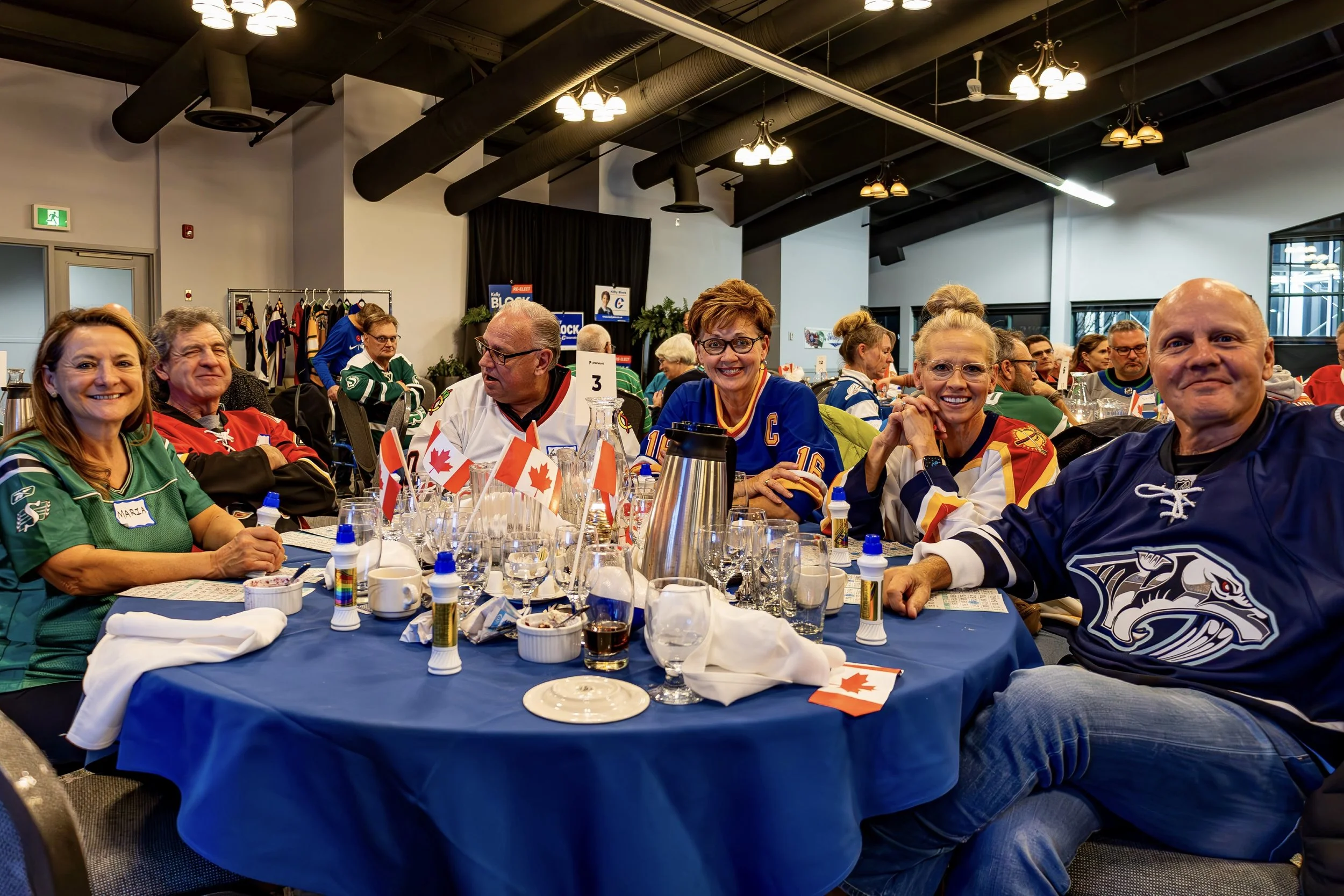Group of people wearing hockey jerseys sitting at a table with Canadian flags during a celebration or gathering indoors.