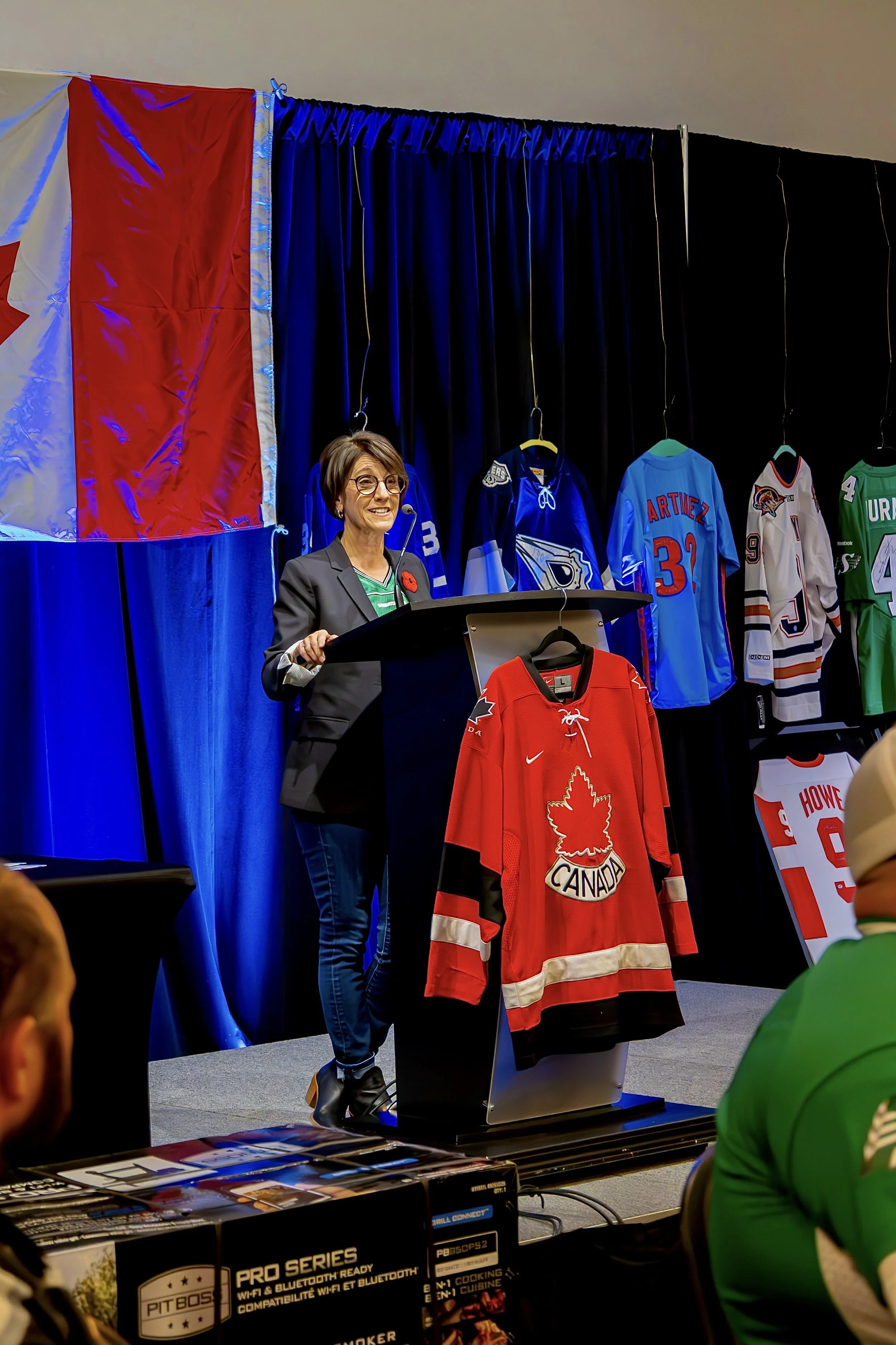A woman with glasses stands at a podium, speaking at an event. Behind her is a display of hockey jerseys and a Canadian flag.