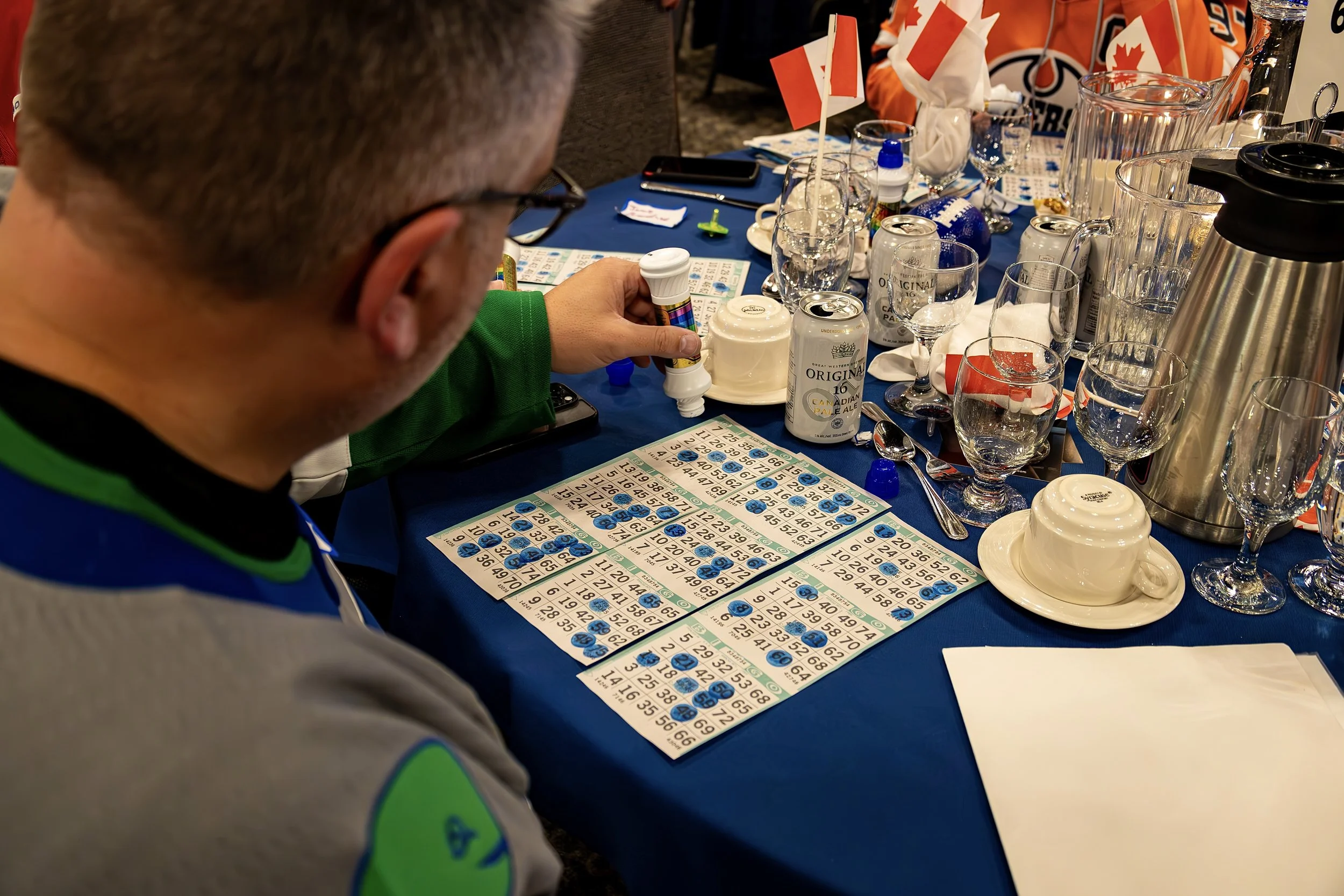 Close-up of a person playing bingo at a table with bingo cards, bottles, glasses, and scattered items on a blue tablecloth.