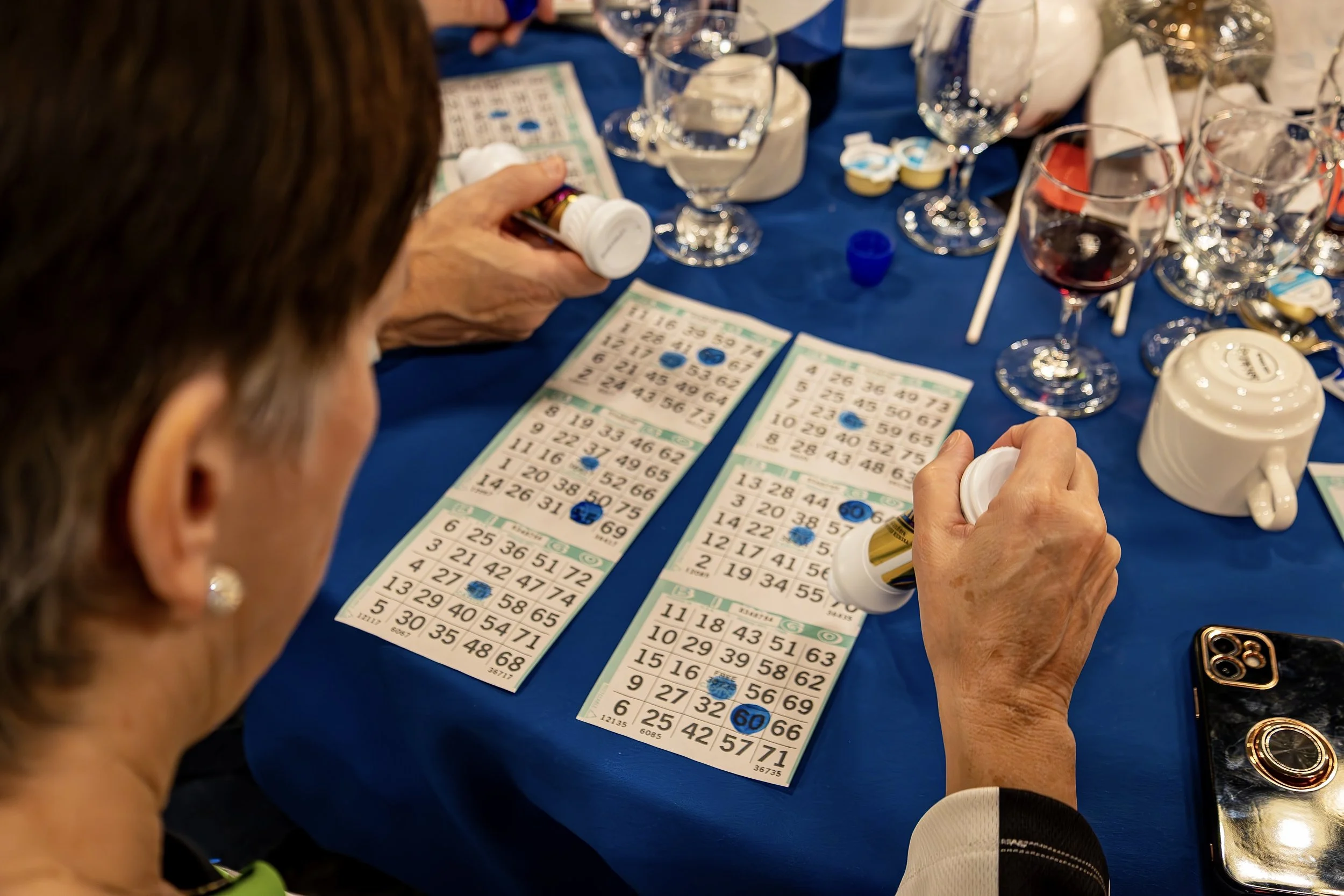 A person playing bingo at a table with bingo cards, markers, glasses of water, a cup, and wine glasses, with folks in the background.