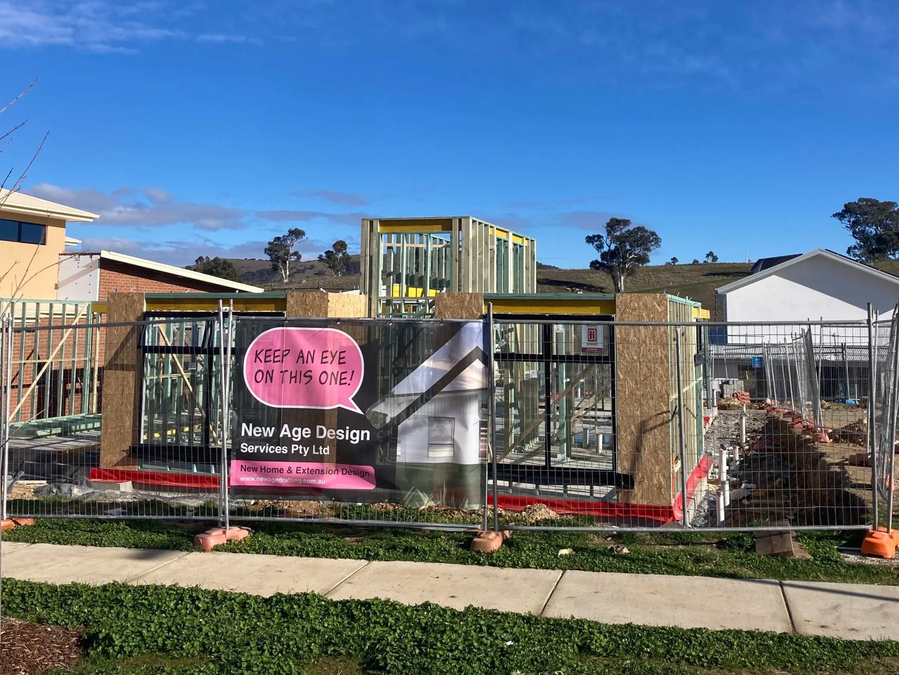 Construction site with a partially built house frame, surrounded by a metal fence, and a sign that reads 'Keep an eye on this one!' indicating new home and extension design services, under a bright blue sky with clouds. New Age Design Project