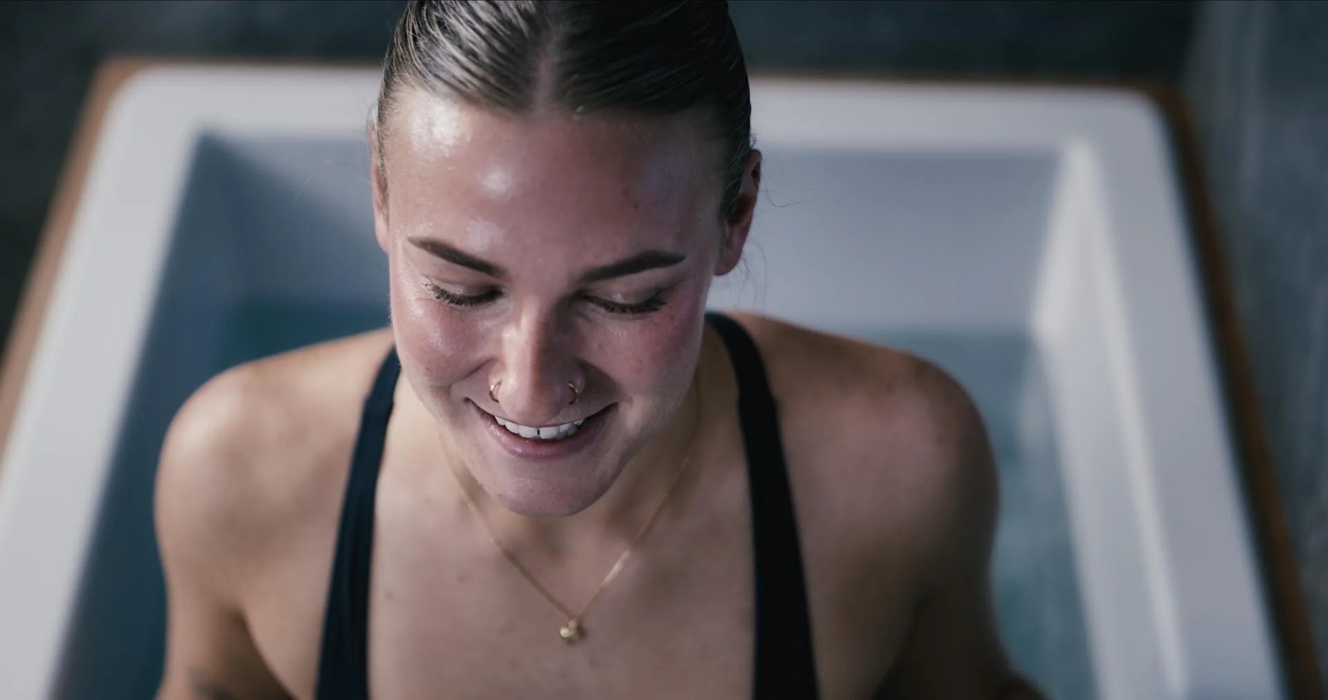 Woman smiling in a ice bath with her head slightly bowed, looking down, hair slicked back, wearing a black tank top and a gold necklace.