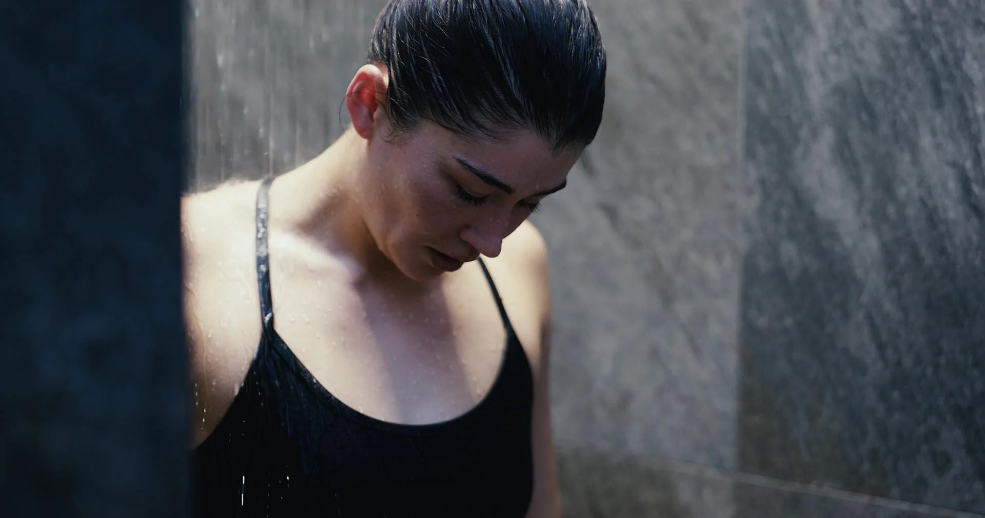 A woman with wet hair and a black tank top looking down, standing under a shower with water falling on her.
