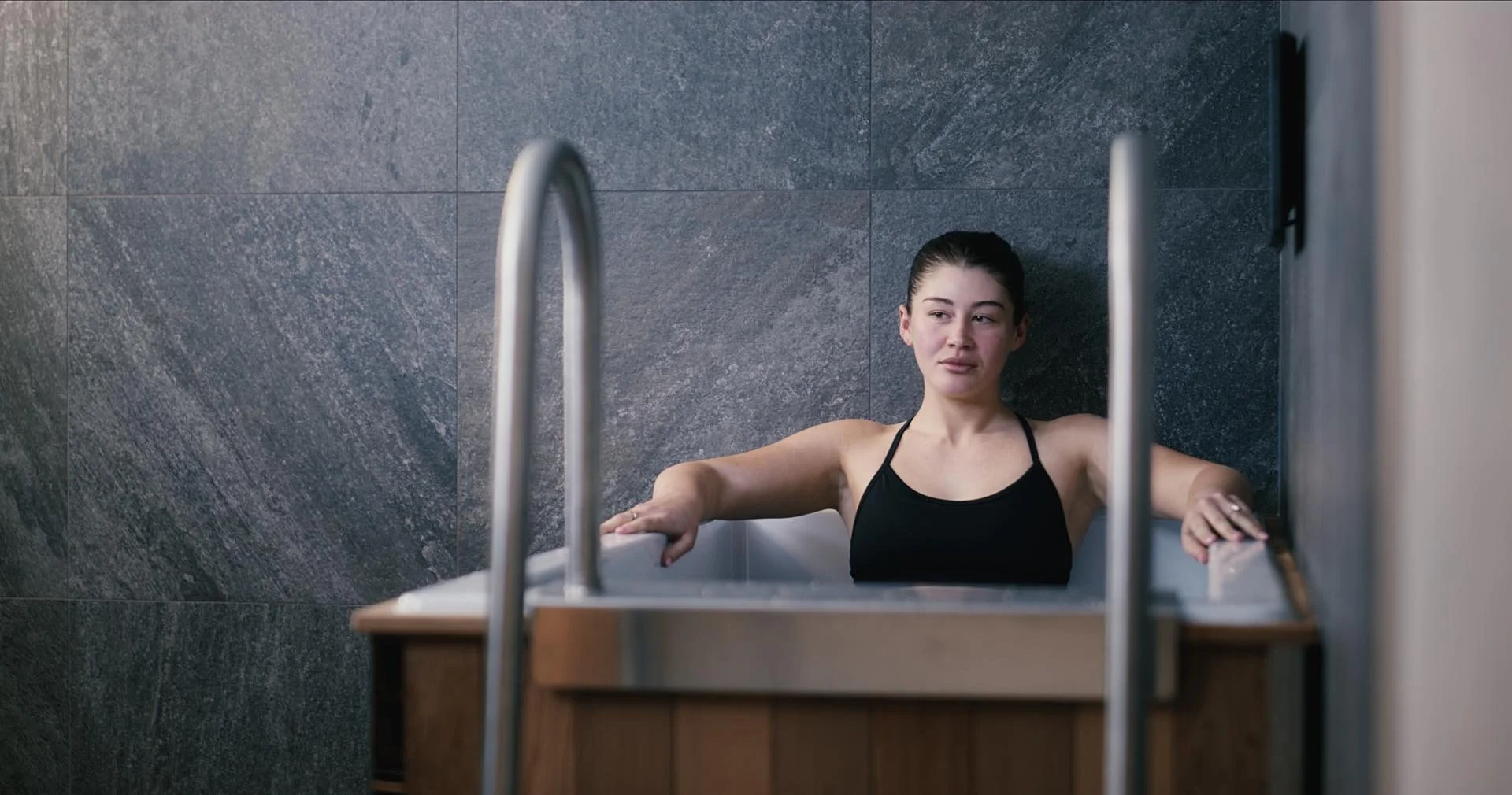 Woman in an emotional state sitting in a ice bath, with a dark gray tiled wall behind her.