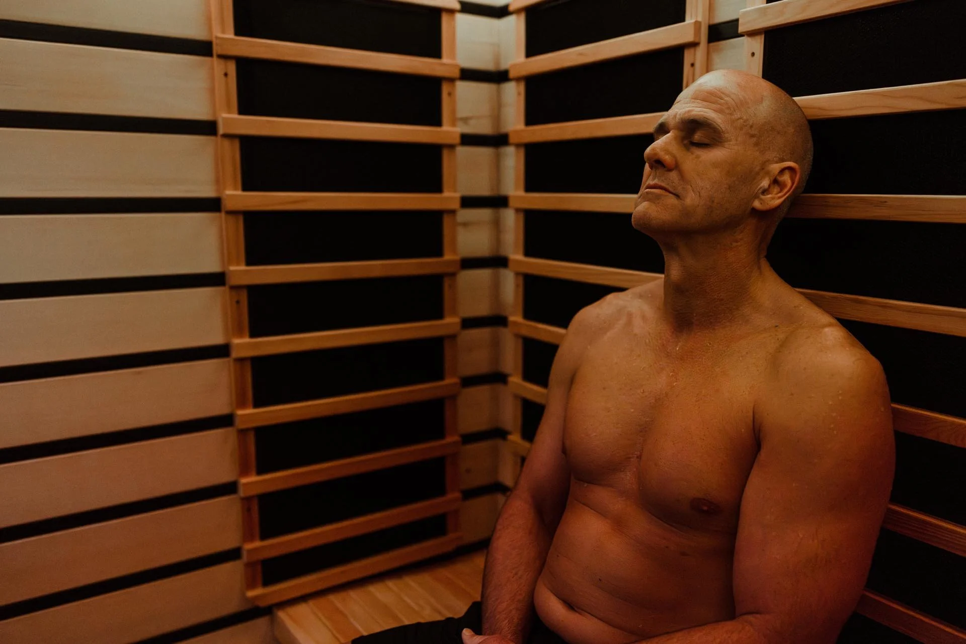 A shirtless man with a shaved head sitting with his eyes closed in a wooden sauna, surrounded by horizontal wood paneling.