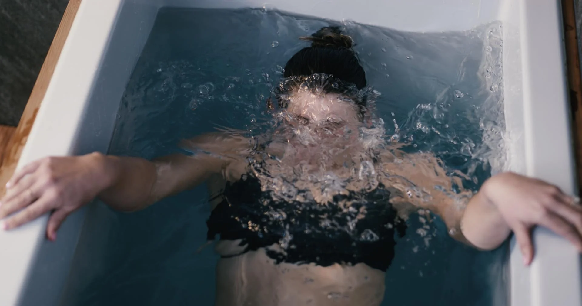 A person submerged in a bathtub, head and shoulders underwater, with arms stretched out holding the sides of the ice bath.