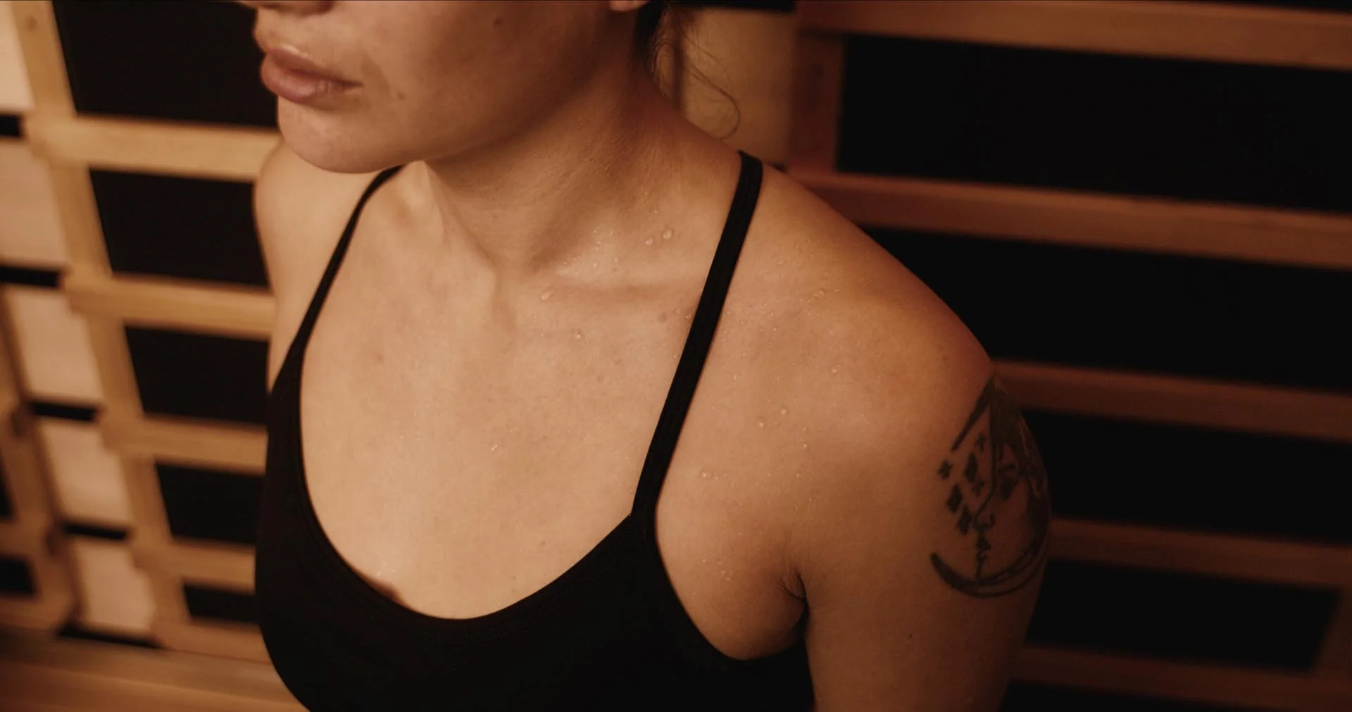A woman with water droplets on her shoulder, wearing a black tank top, with part of a tattoo visible on her upper arm, standing against a wooden background.