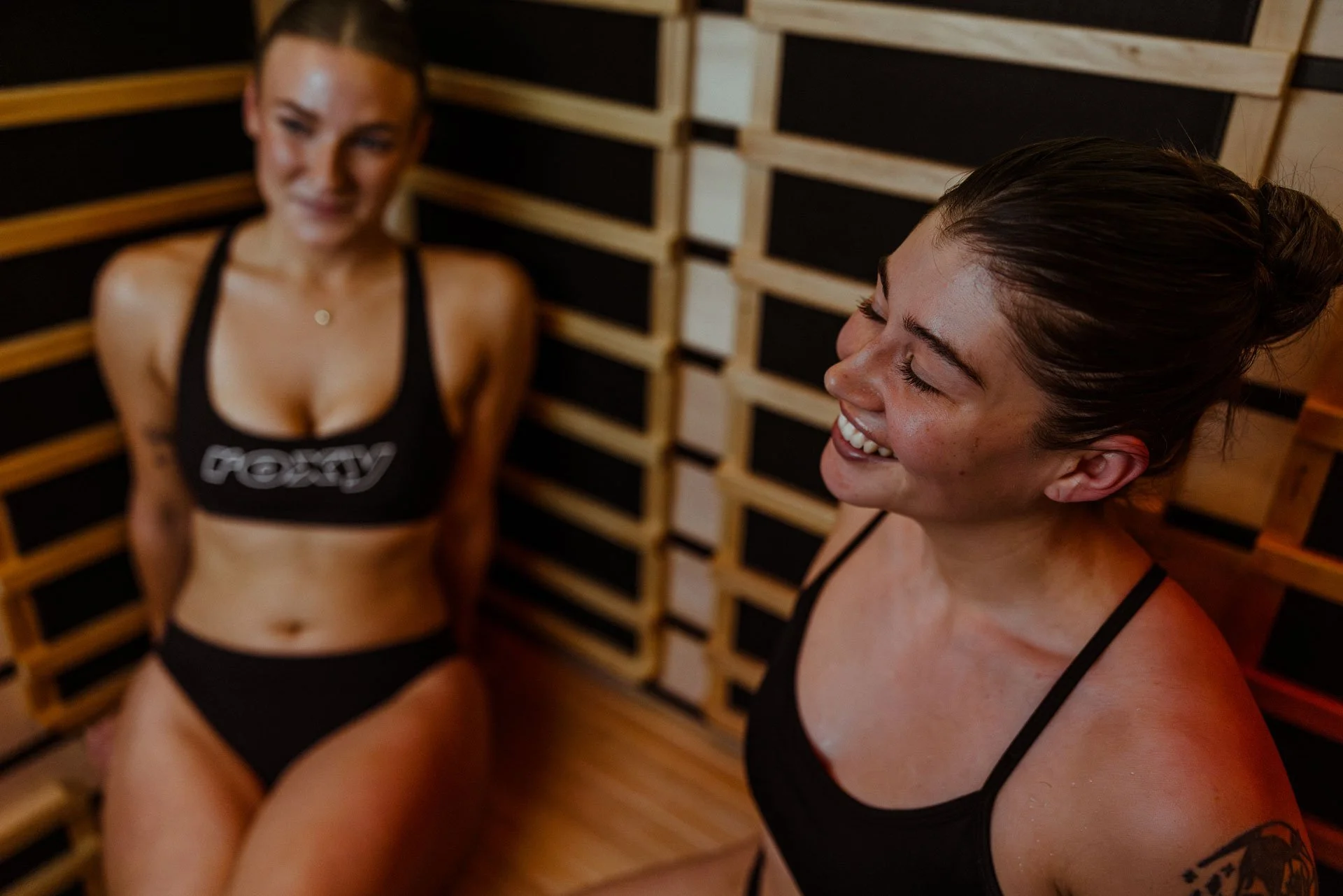 Two women with athletic builds smiling and relaxing in a sauna with wooden paneling.