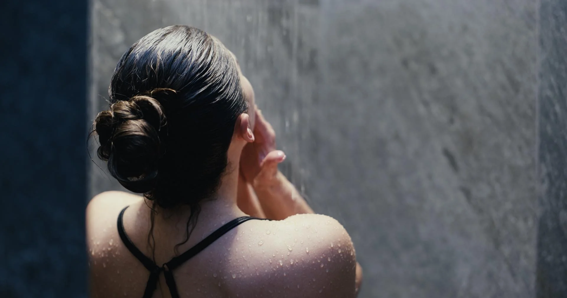 A woman with wet hair in a bun face washing or rinsing her face in the shower.